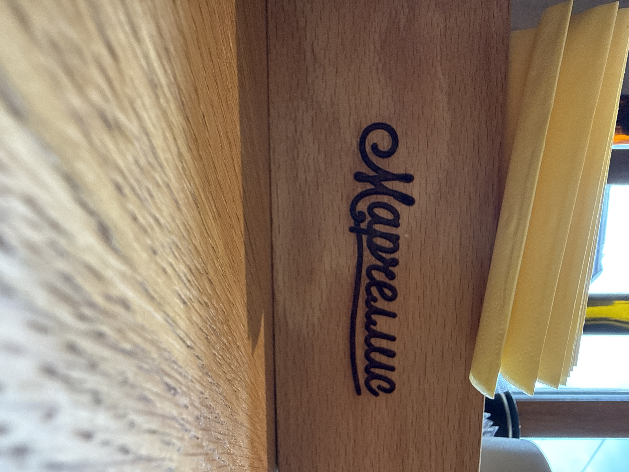 The image shows a wooden box with the Cyrillic inscription "Марселье" in black cursive font. A stack of yellow napkins is resting on top of the box. The background shows a window with a view of the sky and some out-of-focus objects, including what appears to be a yellow bottle or container and a white cup. The foreground is a wooden surface, possibly a table, with a textured grain pattern. The scene is lit by natural light coming from the window, casting shadows on the wooden surface. The image appears to be taken indoors, possibly in a cafe or restaurant setting. There is no indication of the specific location within Gatchina, Russia.
