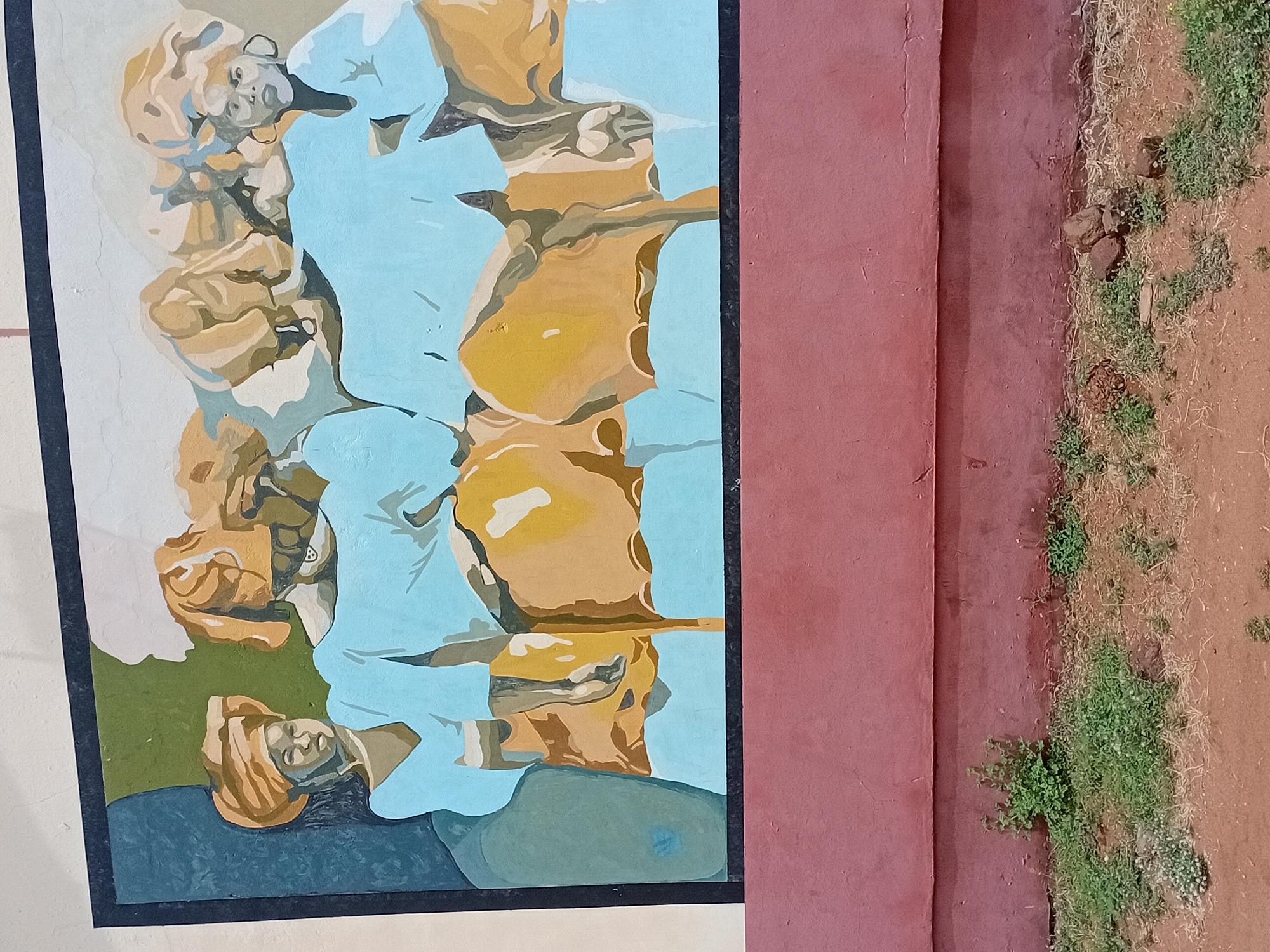 The image shows a painting on a wall. The painting depicts a group of people, with one figure lying down at the bottom and others standing or sitting above. The colors are primarily shades of blue, brown, and tan. The style of the painting appears to be stylized or impressionistic.
The painting is mounted on a wall painted in a reddish-brown color. To the right of the wall, there is dirt and vegetation. Based on the description of Lokoja, Nigeria, I cannot make a detailed description due to the limited image context.