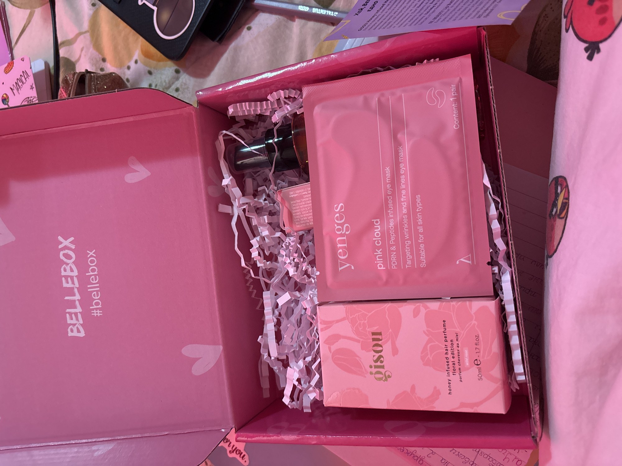 A Bellebox is open, revealing pink-themed beauty products. The box contains Yenges pink cloud PDRN & Peptides infused eye mask, Gisou honey-infused hair perfume, and a small unlabeled bottle. The box is filled with white paper filler. The box is pink with the words "BELLEBOX" and "#bellebox" printed on the side. The product packaging is also pink. The Gisou perfume is the floral edition, parfum cheveux au miel, and contains 50ml - 1.7 fl.oz. The text indicates "Content: 1 pair" for the Yenges eye mask. The box appears to be resting on a patterned surface. There is no indication of Garmen, Bulgaria, in the image.