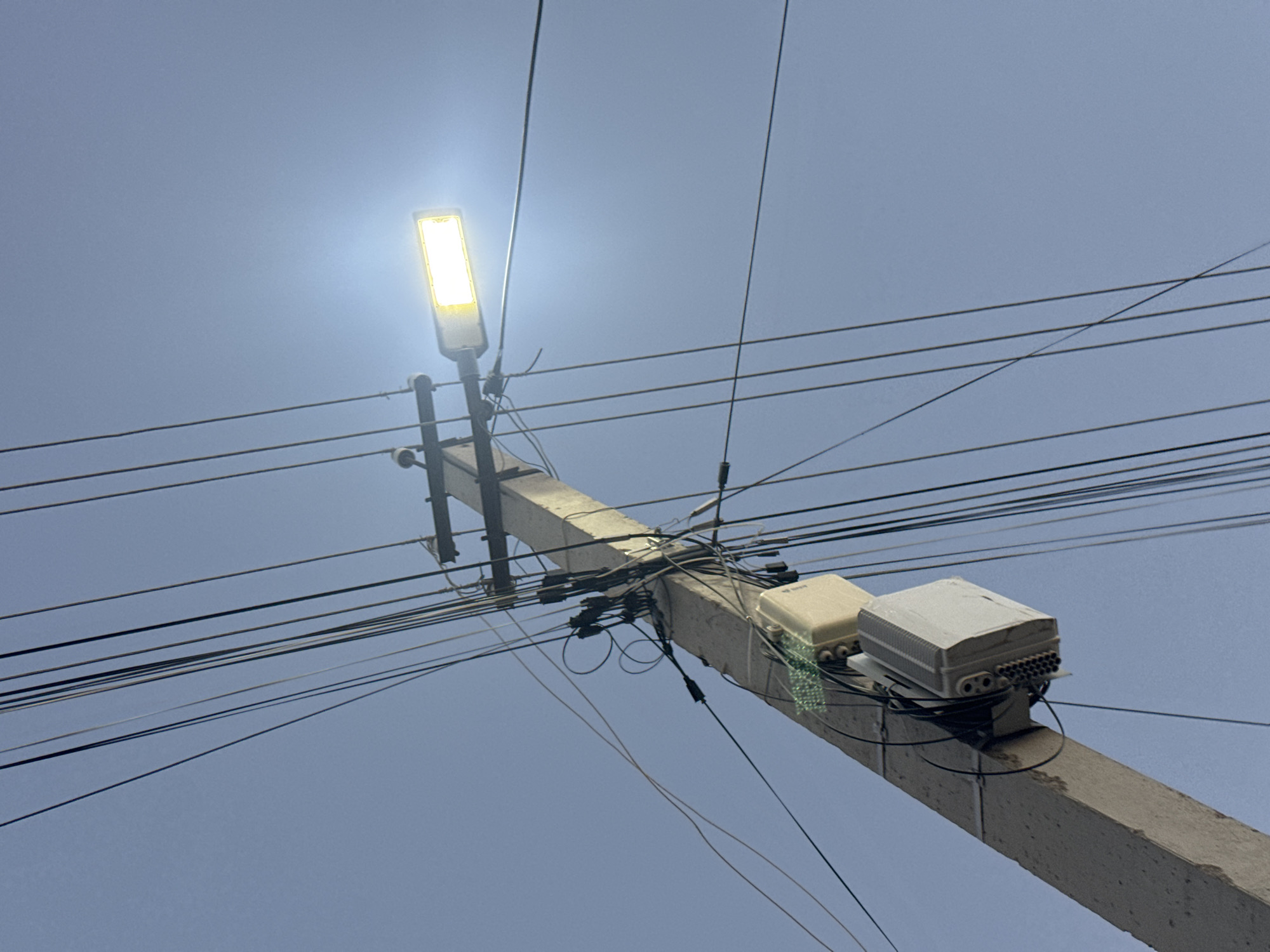 The image shows a concrete utility pole in Atameken, Kazakhstan, against a clear blue sky. Several black wires are strung from the pole, crisscrossing the frame. A modern, rectangular street lamp with a bright yellow-white light is attached to the pole near the top, illuminating a section of the sky. Two beige and white electrical boxes are also mounted on the pole at different heights, one with visible green circuitry. The scene appears to be outdoors, in an urban or suburban environment, focusing on infrastructure.
