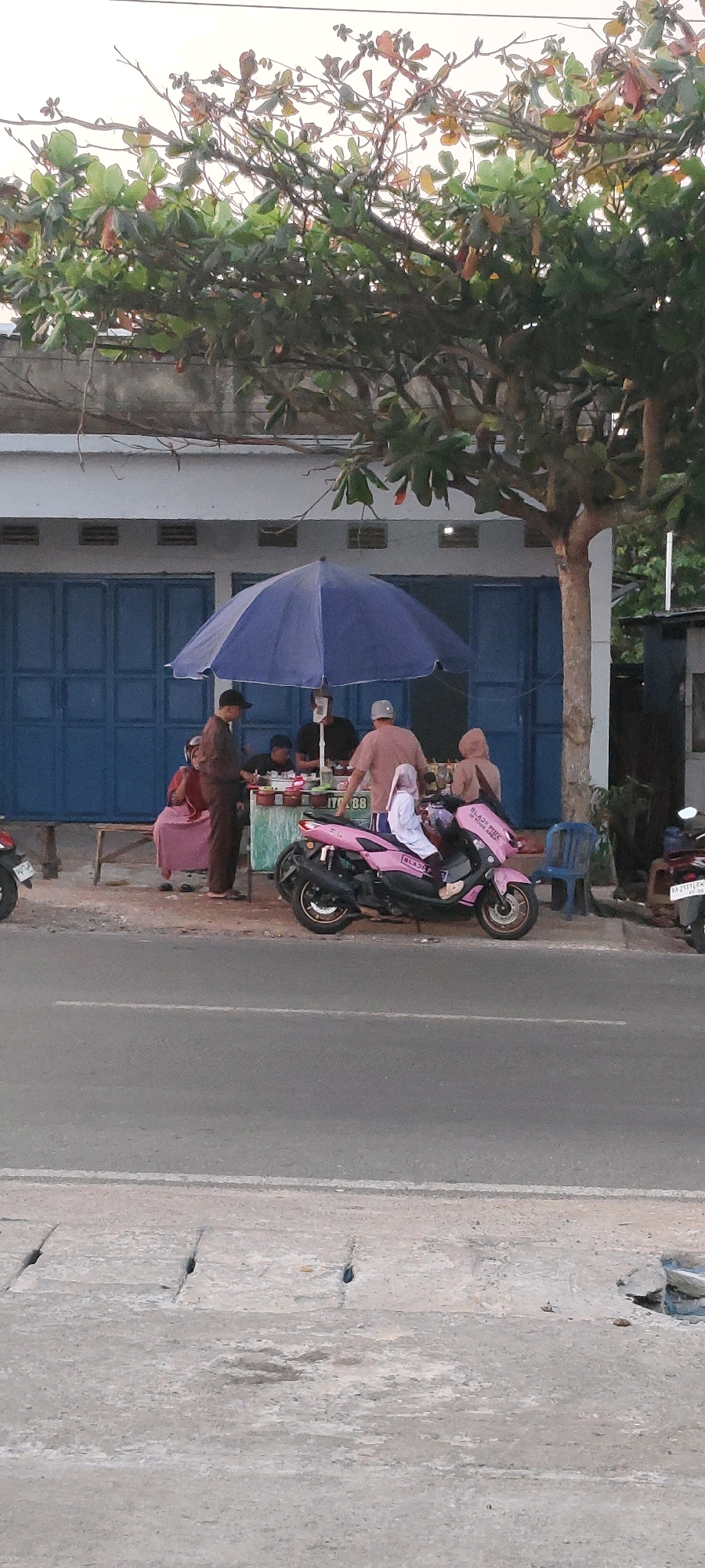 In Bentok Kampung, Indonesia, an outdoor food stall operates under a large blue umbrella. A motorcycle, painted bright pink and adorned with "BLACKPINK" and Korean text, is parked prominently in front of the stall. Three individuals are visible behind the stall, two appearing to be vendors and one customer. A fourth person, wearing a headscarf and white attire, is seated on the pink motorcycle. The stall is set up in front of a building with blue double doors and a light-colored facade. Trees with green and reddish-brown leaves overhang the scene. A paved road is in the foreground, with a concrete drain cover adjacent to it.