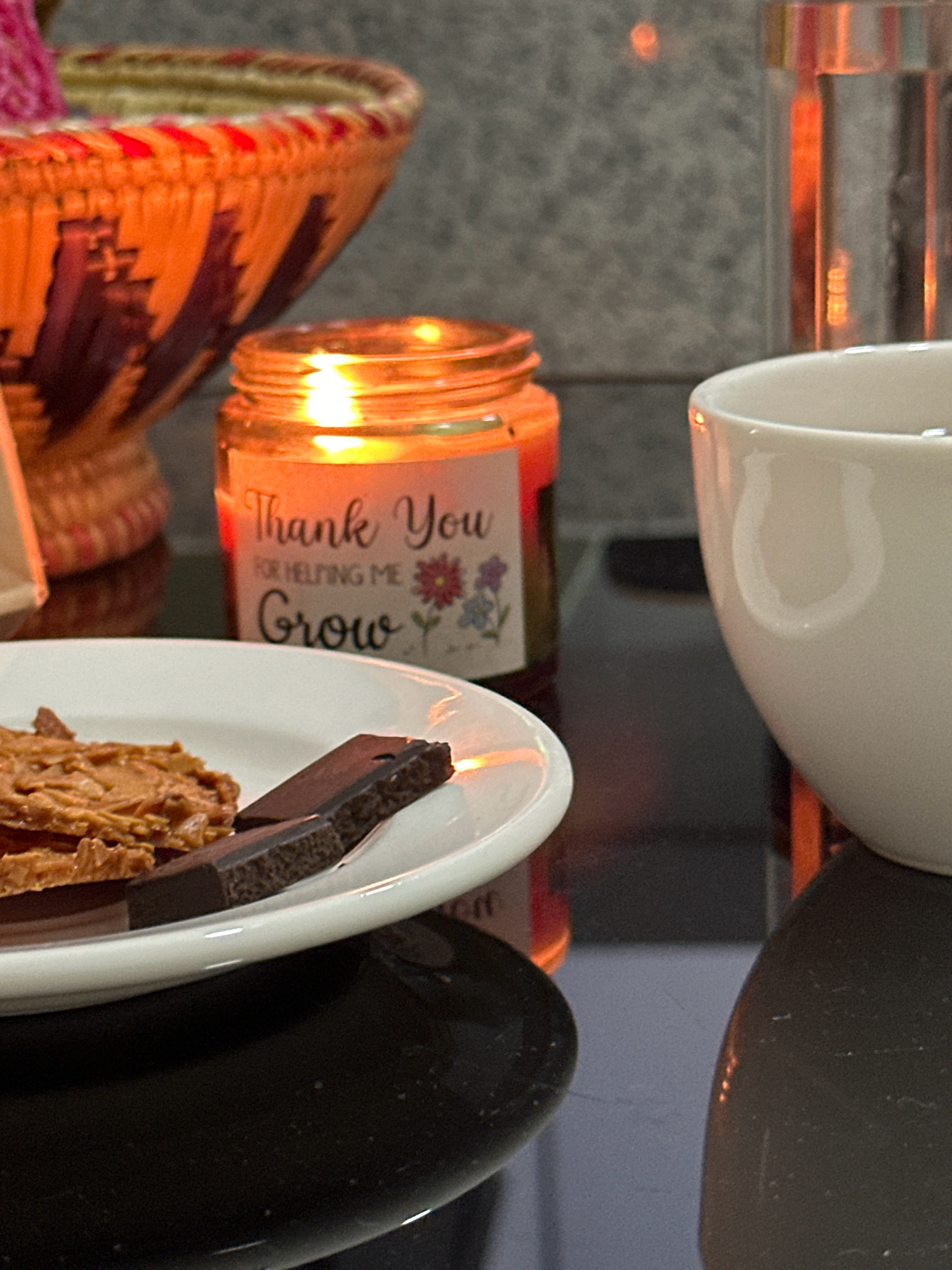 The image shows a close-up of a table with several objects. In the foreground, there is a white plate with some dark chocolate pieces and a textured, cookie-like confection, possibly almond biscotti. To the right of the plate is the rim of a white mug. Behind the plate and mug, a lit candle in a glass jar is visible, with a label that reads "Thank You FOR HELPING ME Grow". The candle flame casts a warm glow. In the background, on the left, is a woven basket with orange and dark red/purple patterns. The surface the objects are on appears to be a dark, reflective material, possibly a table. The background wall is textured, resembling grey stone or concrete. There is no indication of the city of Lalitpur, Nepal.
