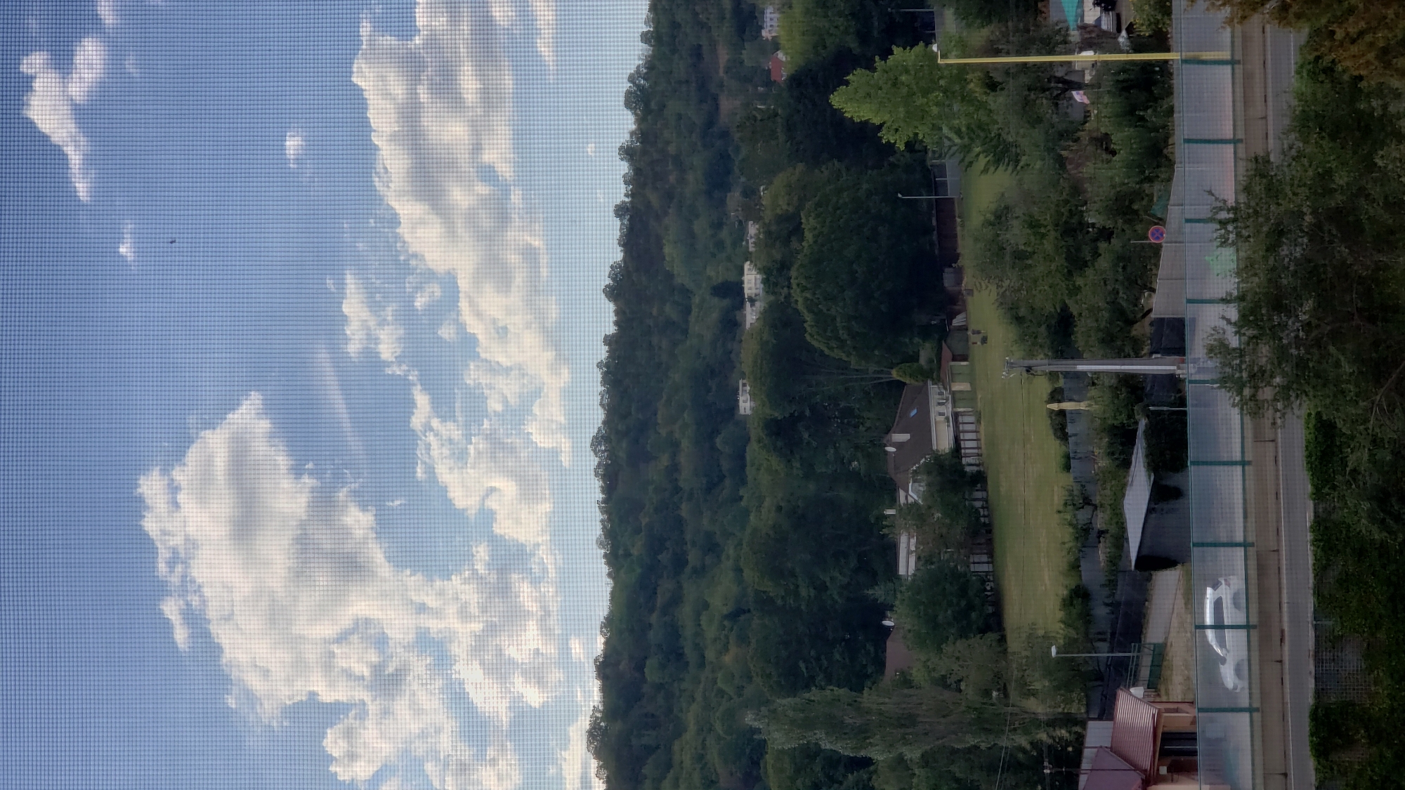 The image depicts a view from a window, likely a residential building, overlooking a suburban or semi-rural landscape. In the foreground, a light-colored car is visible on a road behind a transparent barrier. Adjacent to the road is a building with a red roof and a smaller gray structure. Beyond this, the ground slopes upwards, covered in green grass and scattered trees. Further in the background, a densely wooded hill rises, dotted with a few buildings, including what appear to be apartment blocks or houses, on its upper slopes. The sky is blue with scattered white clouds, and a faint indication of a bird or plane can be seen. The overall impression is that of a residential area at the edge of a natural, forested environment. There is no specific visual information in the image to confirm it is located in Prešov, Slovakia.