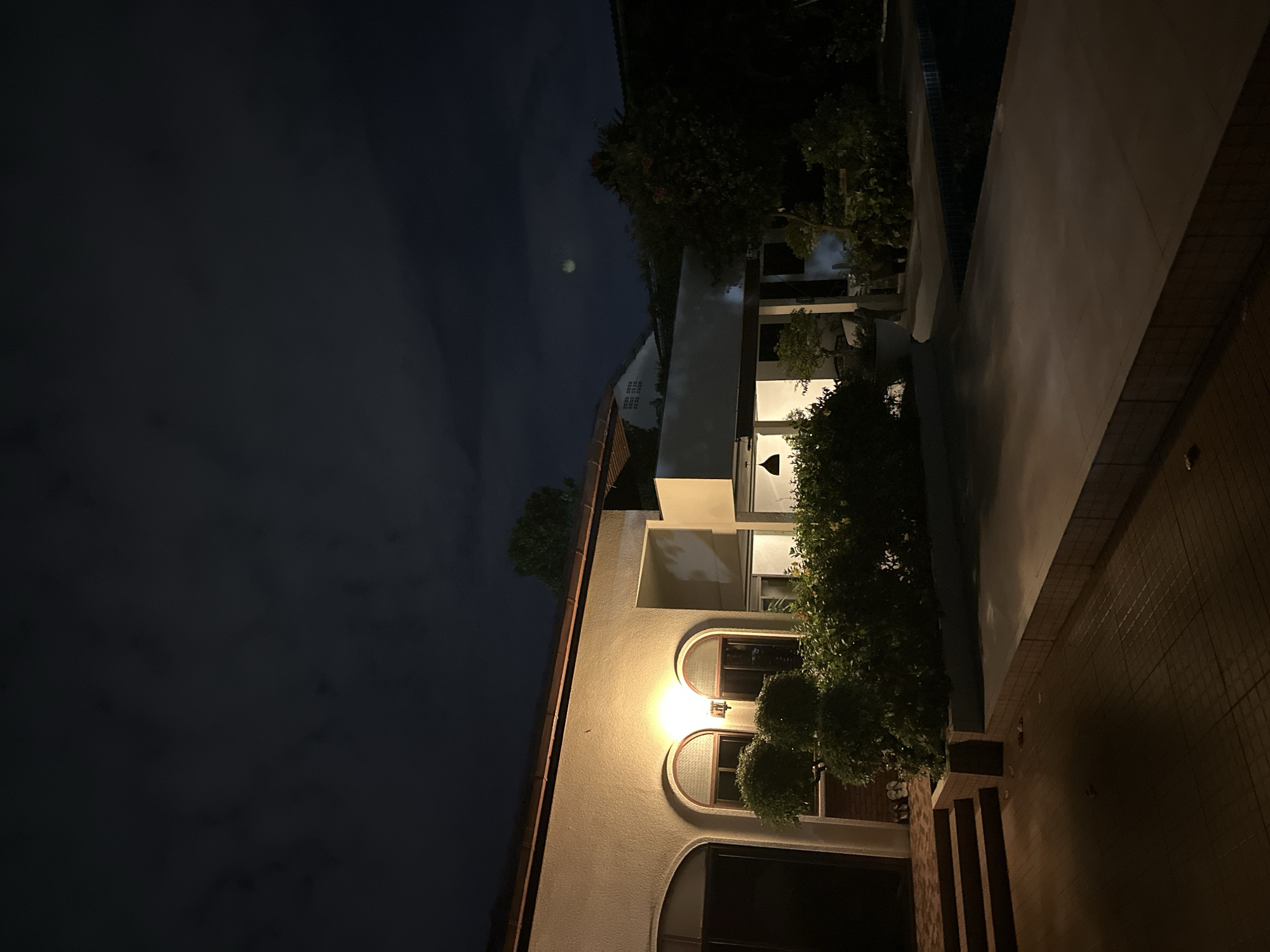 An exterior night shot captures a dimly lit, modern house with a swimming pool. The house features a beige stucco exterior with arched windows illuminated from within. Lush green bushes line the front of the house, and a paved pathway leads to the pool area. The sky above is dark with scattered clouds, and a faint circular object, possibly the moon or a light source, is visible in the upper right portion of the frame. The scene is serene and quiet, with no individuals present.