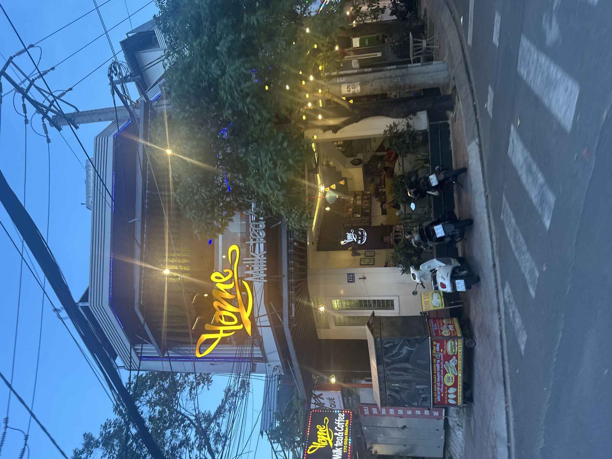 The photograph depicts the exterior of a multi-story building housing the "Home Milk tea & Coffee" establishment, located at 175 Điện Biên Phủ, in the Bac Nha Trang Ward of Nha Trang, Vietnam. The building features a prominent illuminated sign displaying "Home Milk tea" and "Milk tea & Coffee". Additional signage is positioned at ground level, advertising the establishment. Several motorbikes are parked along the curb adjacent to the building, and a food cart is positioned near the entrance. A crosswalk is visible on the street, and overhead power lines are present. The scene appears to be captured at dusk or early evening, as the sky is a deep blue, and artificial lighting illuminates the building and surrounding foliage.