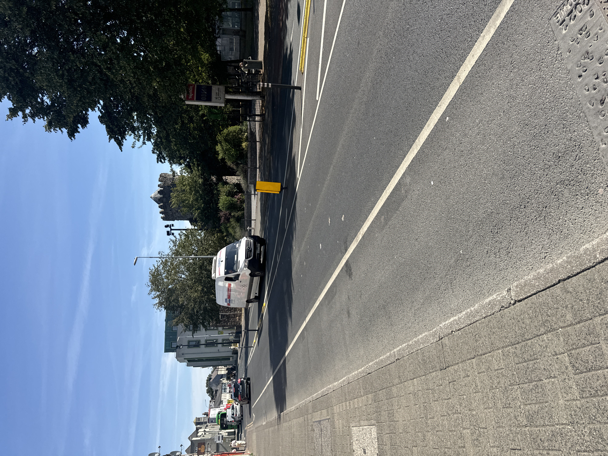 The image depicts a street scene in Waterford, Ireland, likely on a sunny day. The foreground shows a paved sidewalk on the left, made of grey interlocking bricks. The main subject of the image is a road with two lanes, one moving away from the viewer and the other towards. A white van with red and black branding is driving in the lane moving away from the viewer. On the right side of the road, there are trees, a bus stop with a sign, and a glimpse of a stone tower in the distance, possibly a historical landmark like Reginald's Tower. Further down the street, there are buildings, including a modern multi-story building with green accents. Several cars are visible on the road, and a few pedestrians are on the sidewalk. The sky is blue with a few wispy clouds.