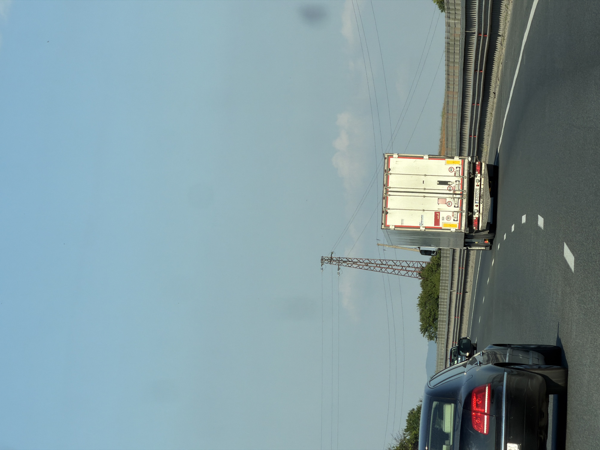 The image shows a sunny day on a highway. In the foreground, a black sedan is driving on the right lane of a multi-lane road. The sedan's brake lights are illuminated. In the middle ground, a white semi-trailer truck is traveling in the same lane as the sedan. The truck has its rear lights on. In the background, a pylon with power lines is visible, and beyond that, trees and hills can be seen. The sky is clear blue with a few white clouds. The highway appears to be in the vicinity of Yana, Bulgaria.