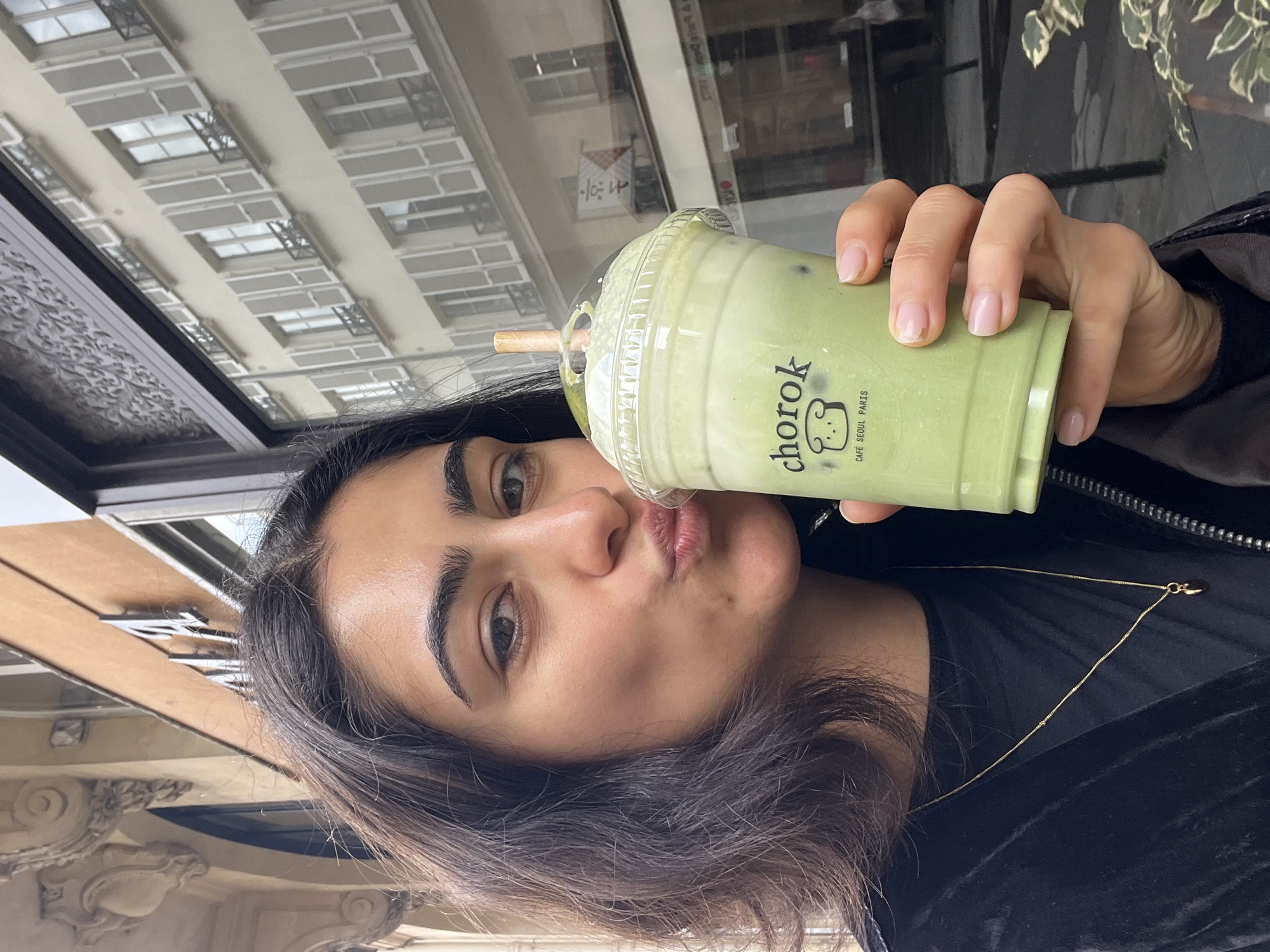 A woman with dark hair, wearing a black shirt and a gold necklace, is holding a green matcha latte with the logo "chorok CAFE SEOUL PARIS" in front of her face. She has her lips pursed as if about to kiss the latte. Behind her, a building facade with multiple windows is visible, reflecting the sky. A window frame and possibly a section of an ornate building are on the left. The location is likely Paris, France, based on the cafe's logo.