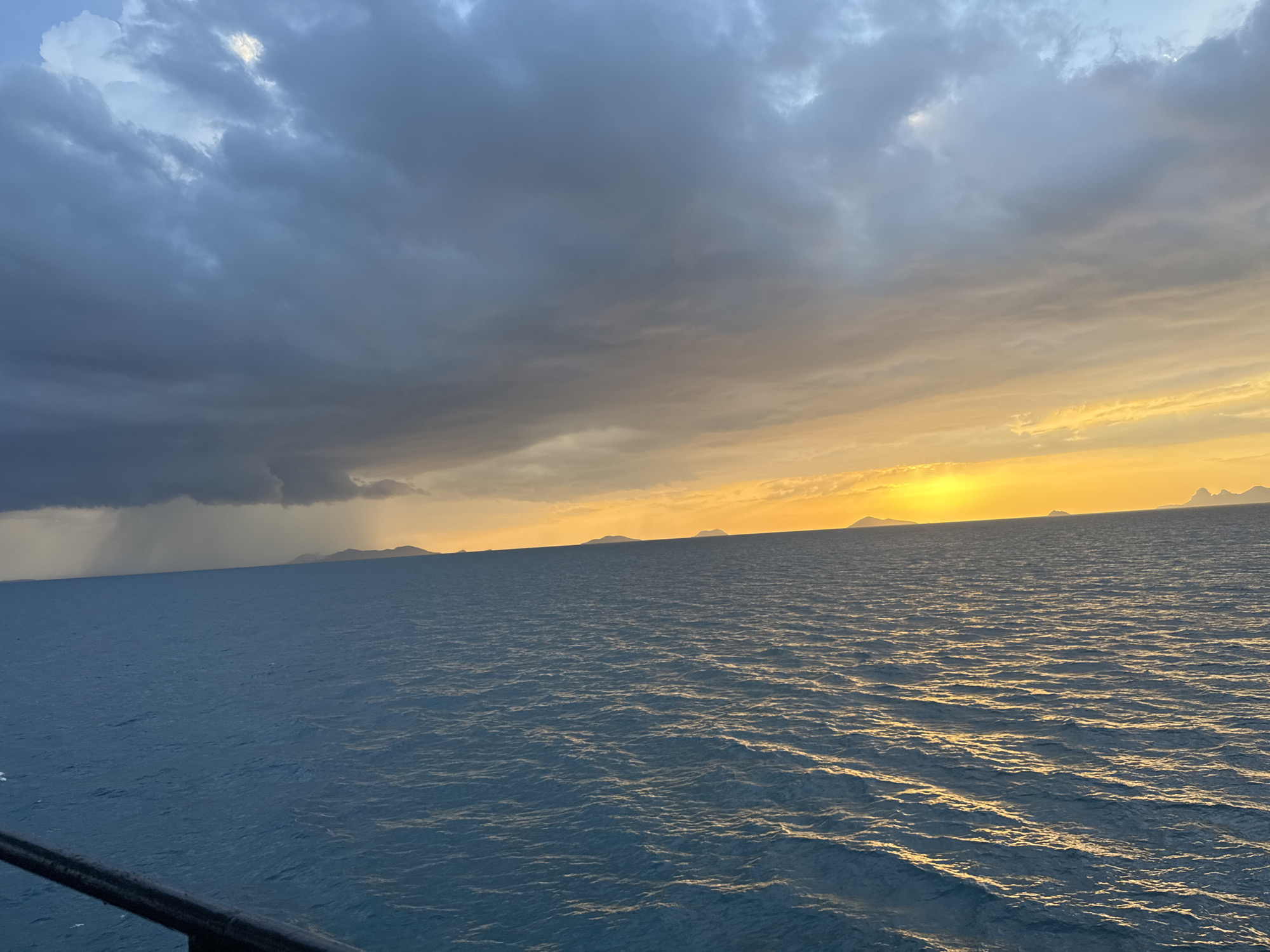 An exterior, eye-level, medium shot captures a dramatic seascape at sunset. The sky is divided, with dark, heavy storm clouds on the left casting rain, contrasting with the bright, golden hues of the setting sun on the right. The sun is partially obscured by clouds, creating a vibrant orange glow that illuminates the horizon. In the distance, silhouetted islands are visible against the colorful sky. The ocean in the foreground is a deep blue, with ripples reflecting the light of the sunset. A black railing or barrier is visible at the bottom of the frame, suggesting the vantage point is from a boat or structure. The scene is likely in Thailand, given the mention of the location, though the specific city is unknown.