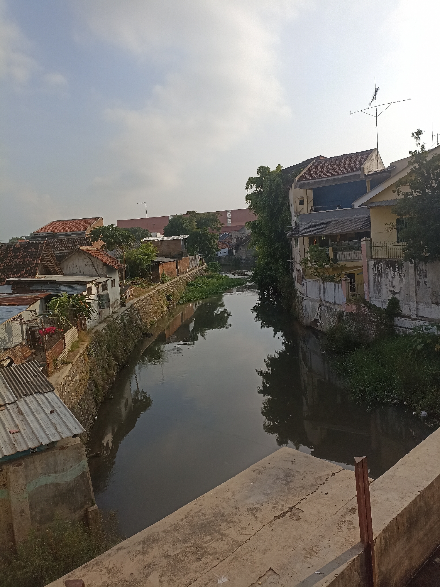 This is a medium-angle shot of a canal in a densely populated area of Banyuwangi, Indonesia. The canal, which runs from the middle ground to the foreground on the right, is dark and reflects the sky and the buildings lining its banks. The water appears calm.
On the left bank of the canal, a row of buildings, some with red tile roofs and others with corrugated metal roofs, are situated close to the water. These structures appear to be residential, with varying degrees of upkeep. A stone wall separates some of these buildings from the canal. There are some trees and vegetation growing along this bank.
The right bank of the canal features several buildings, including a larger, multi-story structure painted yellow with a weathered facade. This building has windows and balconies, and a metal fence in front of it. Trees and lush green vegetation grow along this bank as well.
In the background, more buildings and trees are visible, suggesting a continuous urban environment. The sky above is bright with scattered clouds, indicating it is daytime, possibly morning or late afternoon, given the soft light. The scene depicts a lived-in, somewhat aged urban environment where residential structures are built alongside a waterway.