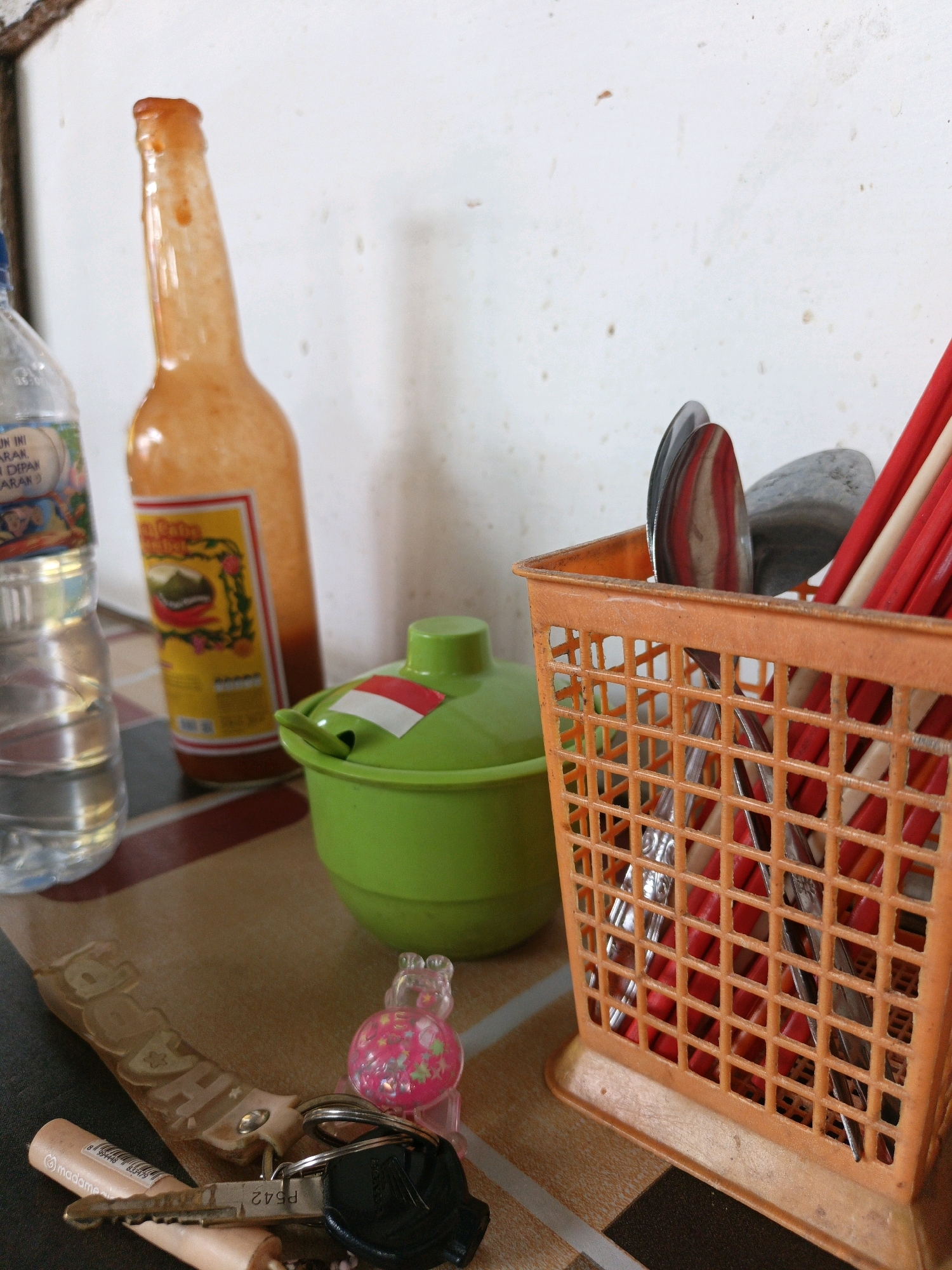 This is a close-up, eye-level shot of a table in Kamulyan, Indonesia, with various items on it. On the left, a clear plastic bottle of water stands next to an orange bottle of sauce with a yellow label. In the center, a bright green bowl with a lid decorated with the Indonesian flag sits on the table. To the right, an orange plastic basket holds chopsticks and spoons. In the foreground, a set of keys with a pink charm and a beige cylindrical object with writing are visible. The table has a tiled surface. The background is a plain white wall.