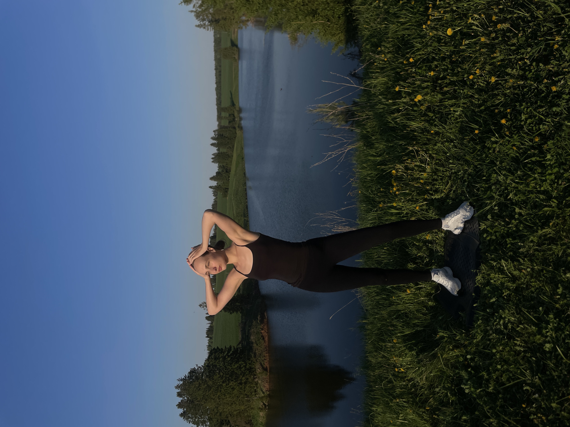 A woman in black athletic wear and white sneakers stands on a grassy area dotted with yellow flowers, facing a body of water, possibly a river or lake, in Подшивалово, Russia. She is holding her hands on her head. Trees line the opposite shore of the water, and a clear blue sky is visible in the background.