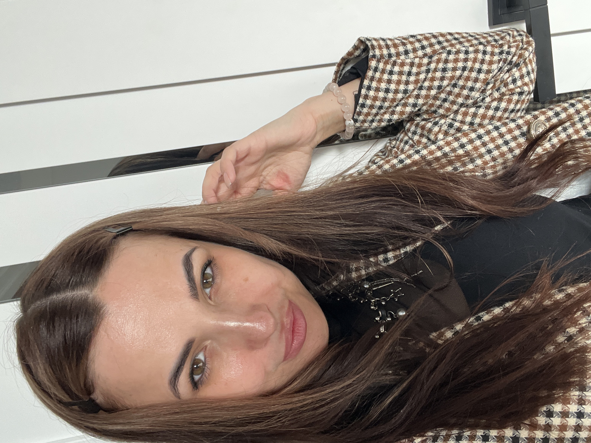 A woman with brown hair and green eyes is looking at the camera. She is wearing a black shirt and a brown and white houndstooth jacket. She has a bracelet on her left wrist. The background is a white wall with a black accent. The location is Unknown, Unknown.