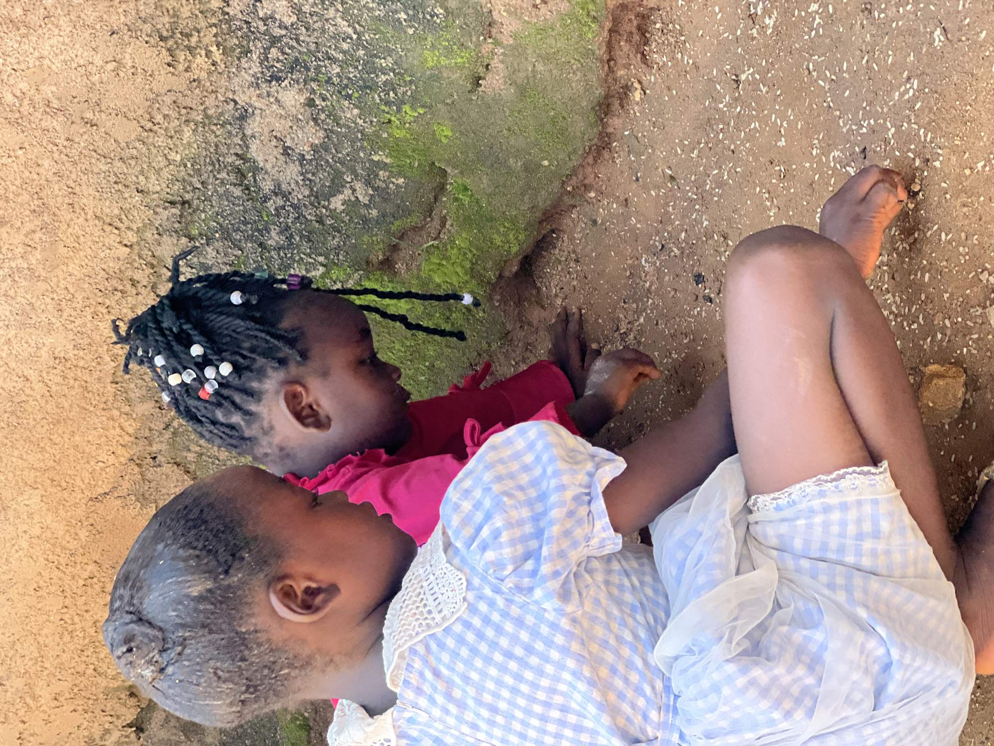 Two young girls are crouched on the dirt ground, leaning forward as if examining something. The girl on the left wears a light blue and white checkered dress with a white lace collar. Her hair is pulled back into a bun. The girl on the right is wearing a bright pink top and her dark hair is braided with small white and colorful beads. She has her head down, focused on the ground. The ground is sandy and speckled with small white grains, possibly rice. Behind them is a rough, textured wall that appears to be made of concrete or mud brick, with patches of green moss growing on it. The scene suggests an outdoor setting in a casual, possibly residential area in Jos, Nigeria.