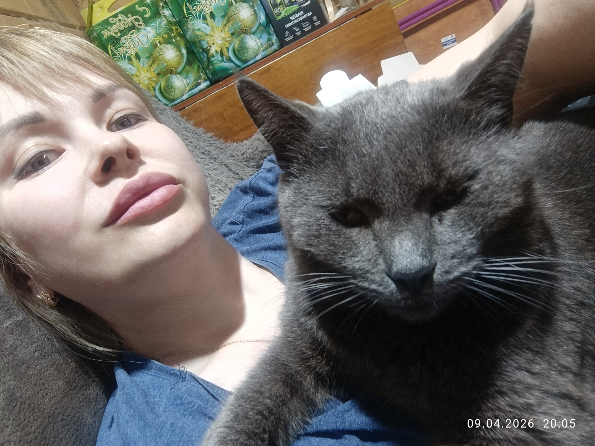 A woman is taking a selfie with a grey cat. The woman has blonde hair and is wearing a blue denim shirt. The cat is mostly grey, with long white whiskers. Behind the woman are several green and gold boxes. The date 09.04.2026 20:05 is printed in the bottom right corner. The location is not discernible based on the available visual information, so the inclusion of Chaikovsky, Russia is not possible.