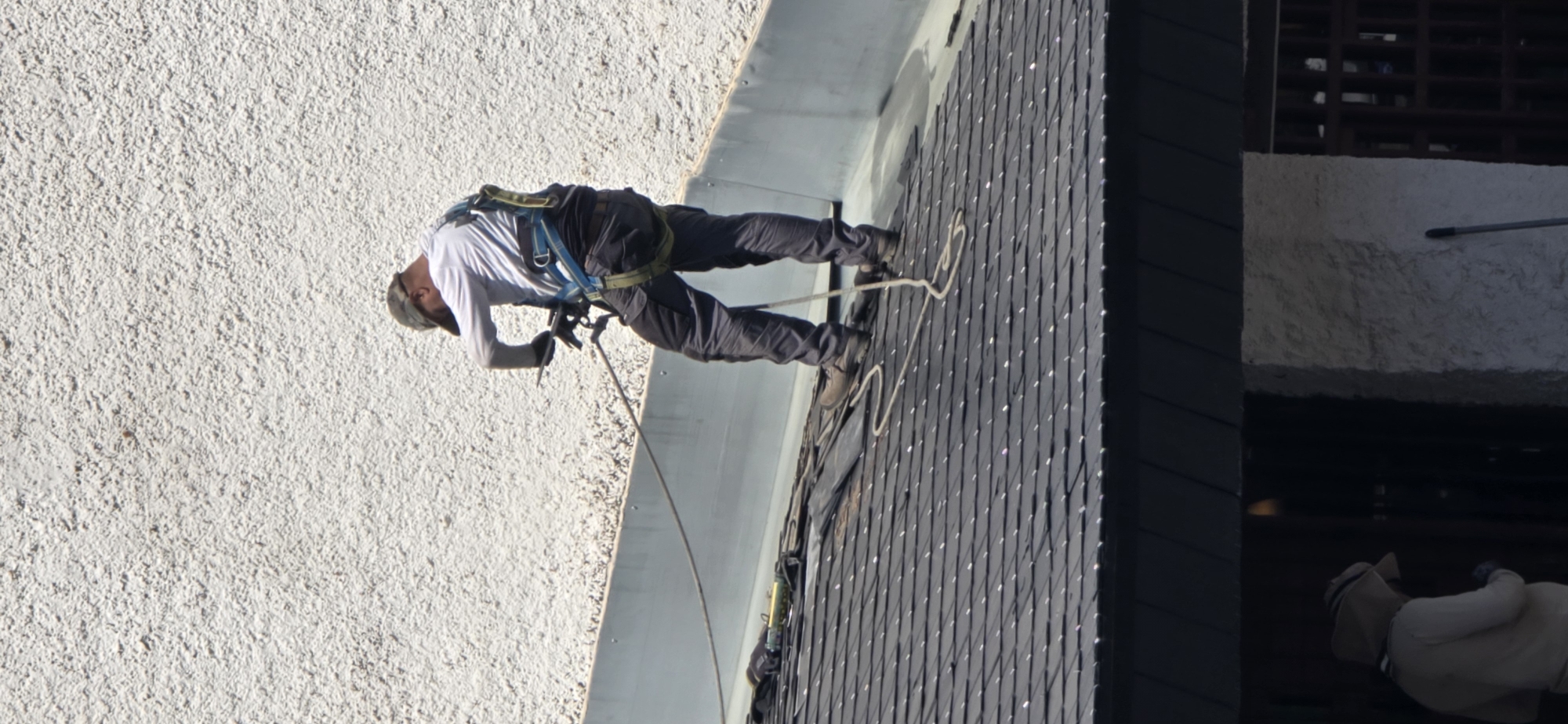 A person is rappelling down a roof on a sunny day in Sallent de Gállego, Spain. The person is wearing a harness and safety equipment, and is holding a tool in their hand. They are standing on a dark-colored, shingled roof. A white building with a textured wall is visible behind the person. The lower part of the image shows a dark overhang of a building, with a person in a hooded garment standing in an entryway.