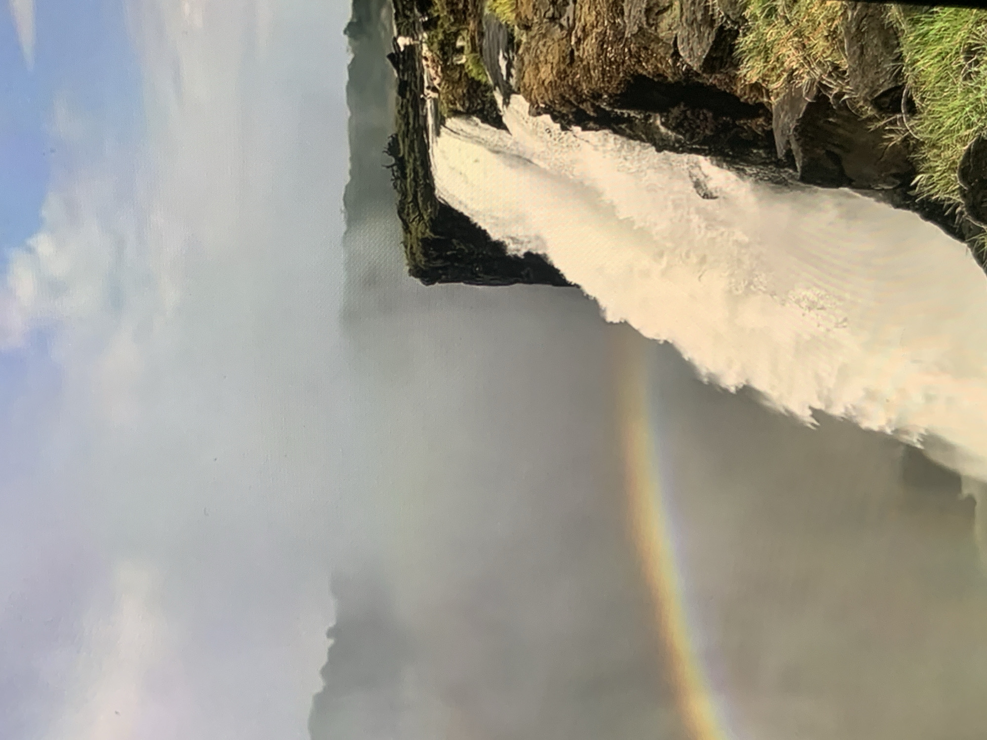 The image shows a powerful waterfall cascading over a rocky cliff. The mist generated by the waterfall creates a hazy atmosphere, and a rainbow is visible in the lower left portion of the frame. The top of the cliff is covered in vegetation, and several people can be seen standing near the edge, observing the falls. The background consists of lush green foliage and a partly cloudy sky. The image does not depict the city of Stavropol, Russia, or any location within it.
