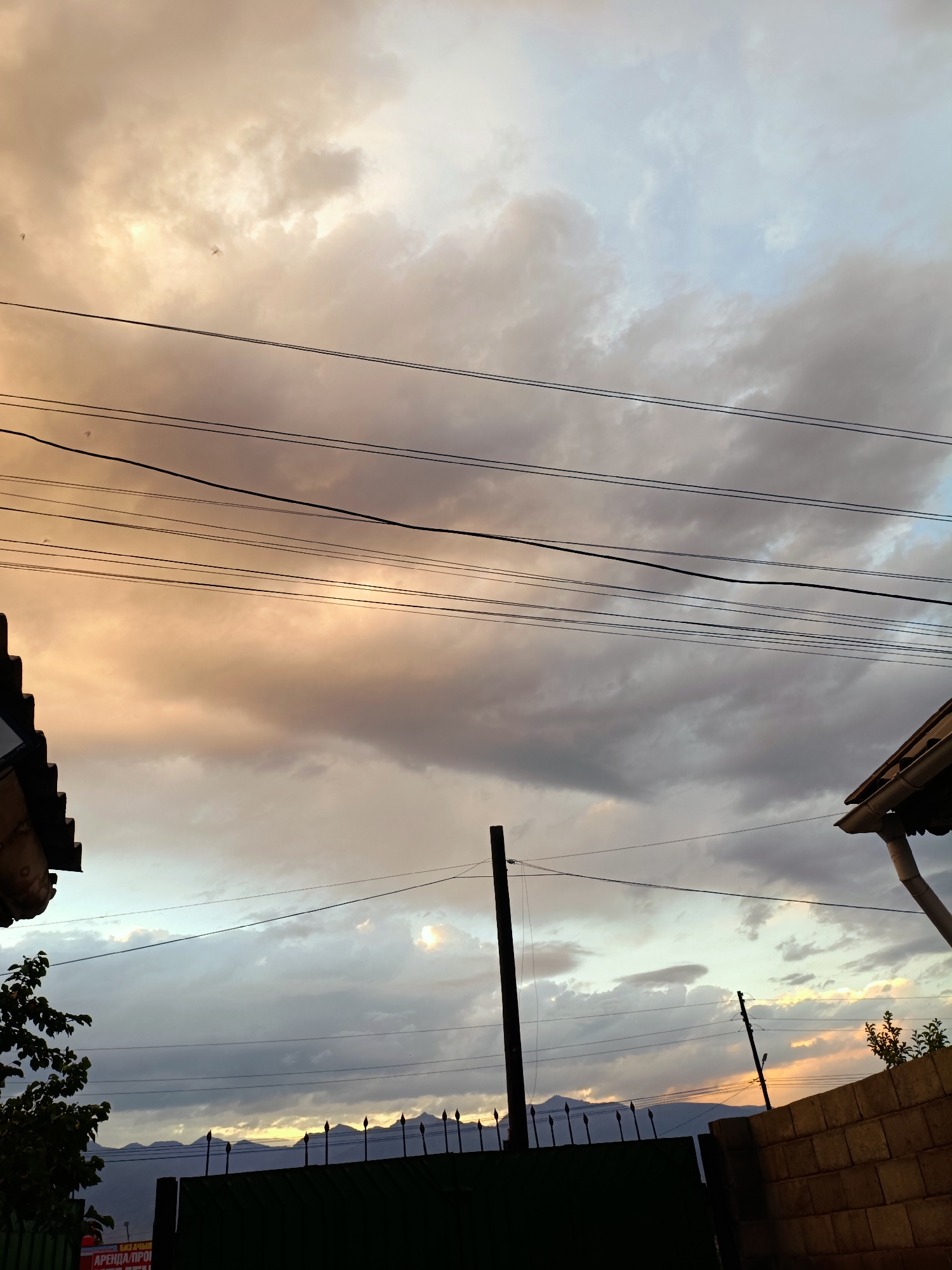 The image depicts a cloudy sky at sunset with several power lines crisscrossing the frame. The sky exhibits a gradient of colors, from warm orange and yellow hues near the horizon to cooler blues and grays higher up. In the foreground, parts of buildings with dark roofs are visible on the left and right edges of the image. A dark fence with pointed finials runs across the lower middle section, partially obscuring a distant mountain range. A silhouette of a tree branch is visible on the left. The scene appears to be an outdoor environment within a residential or semi-rural area.