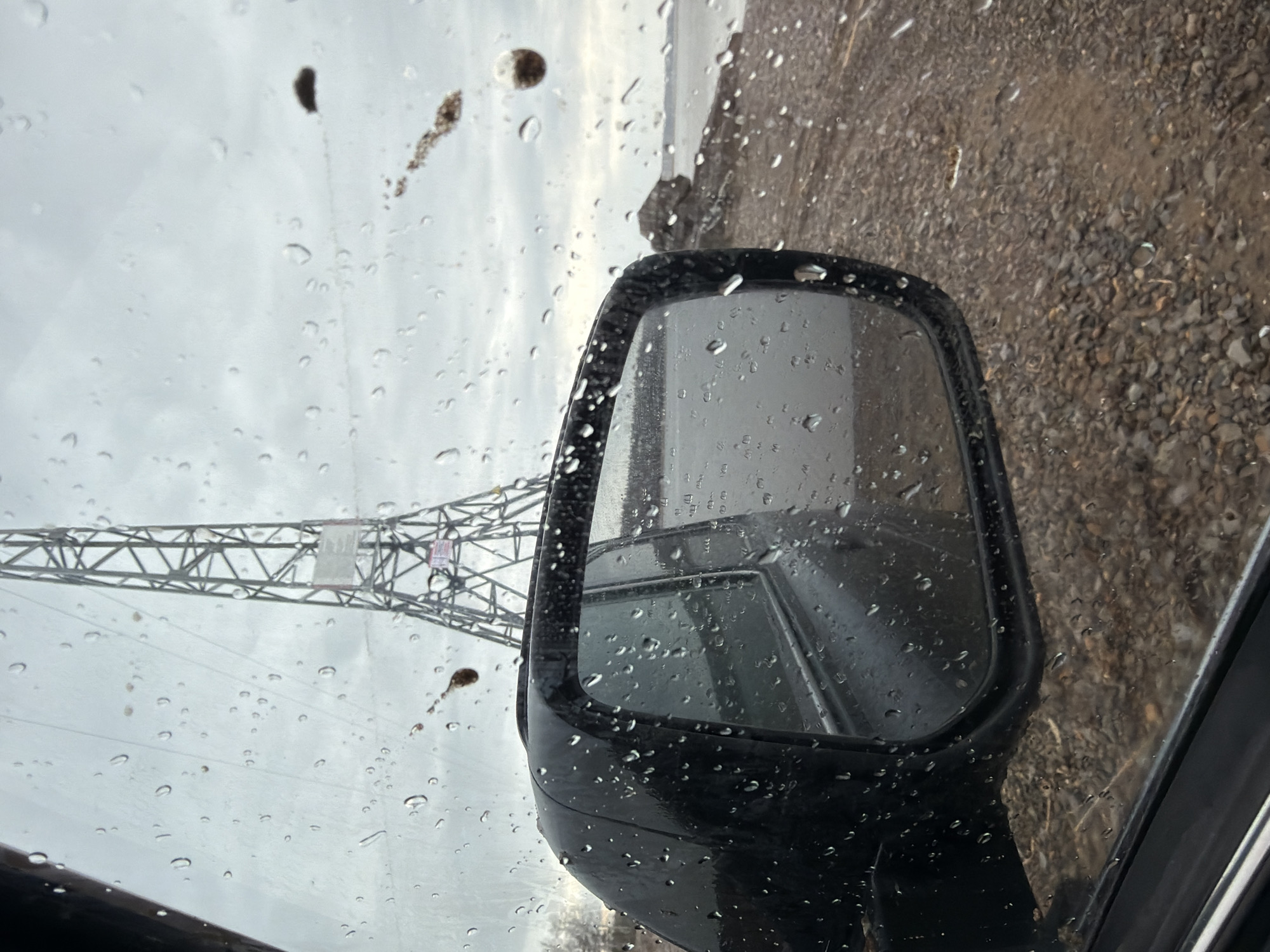 The image shows a view from inside a vehicle on a rainy day. The car's side mirror is prominently featured in the foreground, covered in raindrops. The mirror reflects the interior of the car and the environment around it. The ground appears to be gravel or dirt, also wet from the rain. In the background, visible through the rain-streaked window, is a power transmission tower. The sky is overcast and gray. There is no information that would allow the identification of this location as Chaykovsky, Russia. There are no people visible.
