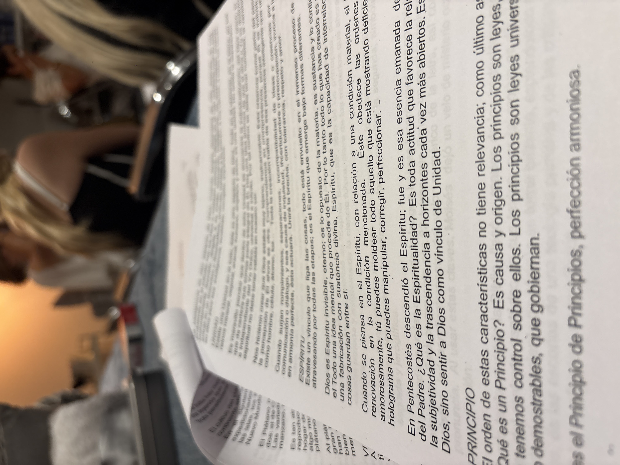 The image shows a close-up of a printed document, likely a book or pamphlet, with text in Spanish. The text discusses concepts related to spirit, spirituality, and principles, seemingly from a religious or philosophical perspective.
The document is printed on white paper with clear, legible text. The text appears to be about spiritual principles, relating to spirituality.
The image's environment hints at an indoor setting, likely a home or office. A person with blonde hair is partially visible in the background, sitting in a chair. There are indistinct objects on a table, possibly indicating a casual setting. The scene lacks specific details that would pinpoint the location to Puebla, Mexico, or any other specific place.