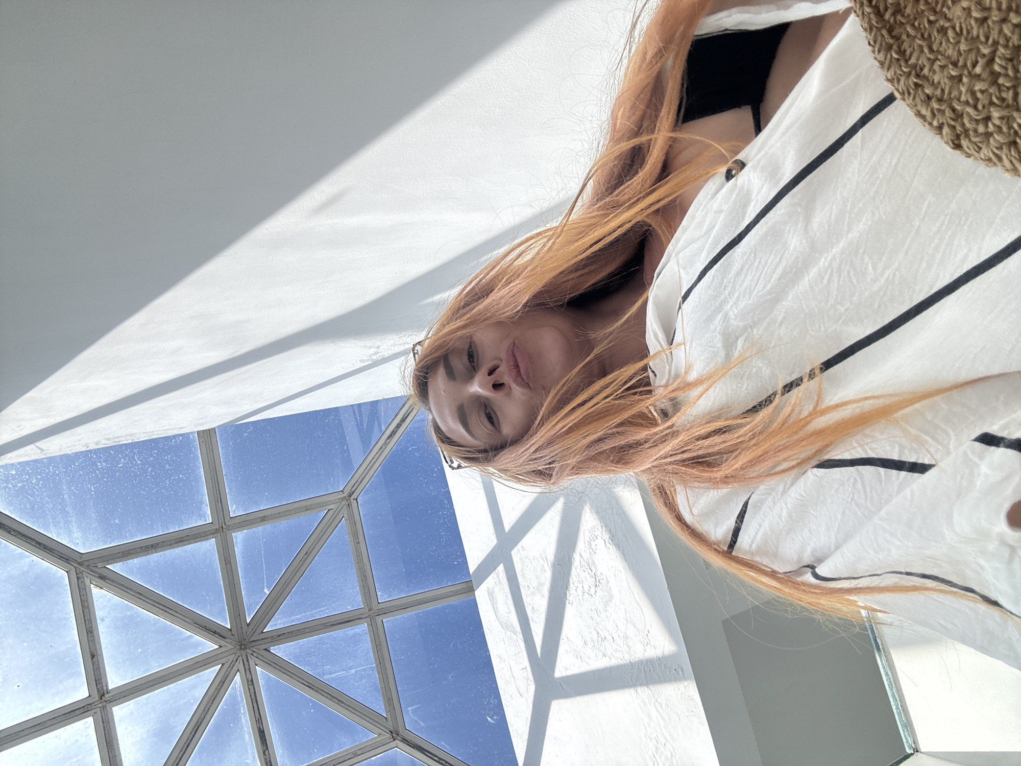 The image shows a woman with long, reddish-blonde hair looking up at a geometric glass ceiling. She is wearing a white shirt with black stripes and a black bikini top underneath. The ceiling is part of a modern building with white walls and natural light streaming in from the sky. The perspective suggests the photo was taken from a low angle, looking upwards. While there are no specific landmarks visible in the image to definitively place it in Budva, Montenegro, the architecture with the open, airy feel and the bright natural light are consistent with designs found in many coastal cities, including Budva, which is known for its modern hotels and resorts. The image captures a moment of casual relaxation.