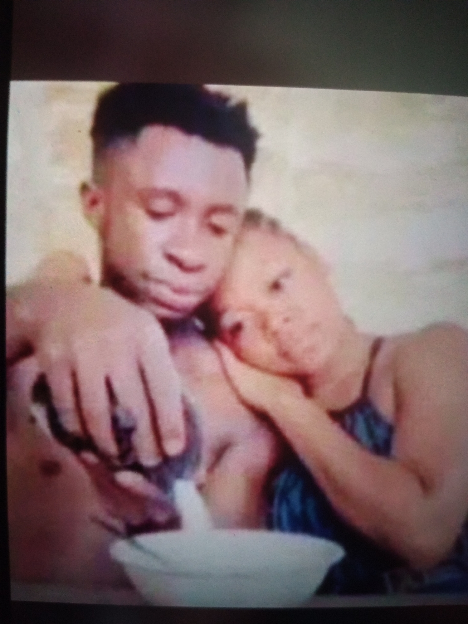 A close-up, low-resolution image depicts a couple embracing. The male, on the left, has short, dark hair and is pouring a white liquid from a dark container into a white bowl. His left arm is around the female. The female, on the right, leans her head on the male's shoulder, her eyes closed. She has light-colored skin and dark hair styled in braids. The background is a plain, light-colored wall, suggesting an indoor setting, possibly a home or studio in Jalingo, Nigeria. The image quality is poor, with visible digital noise and pixelation, making precise identification of facial features and finer details challenging.