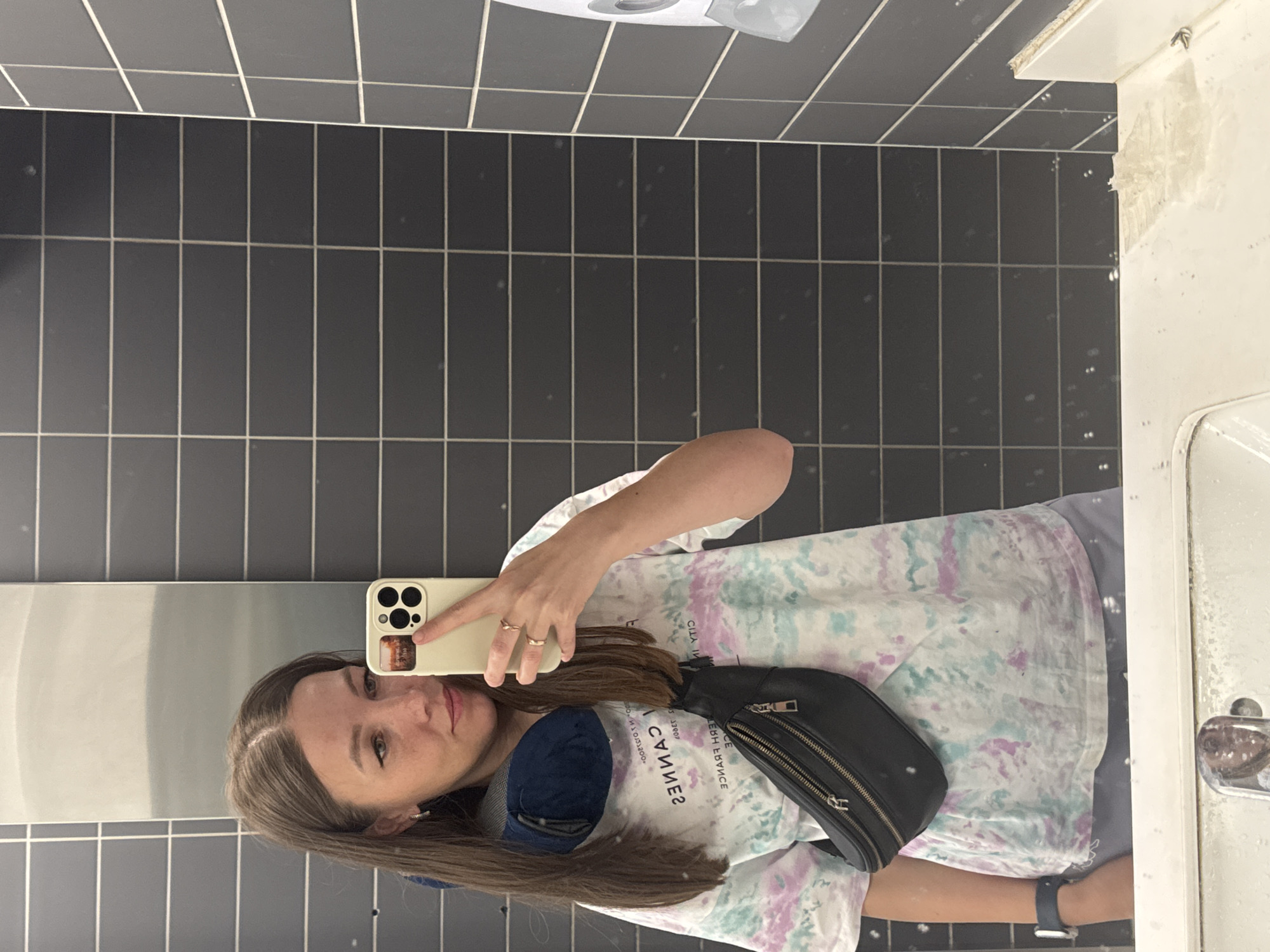 A woman takes a selfie in a public restroom. The background features black, rectangular tiles covering the wall. The woman wears a tie-dye t-shirt, a blue face mask around her neck, and a black fanny pack across her body. She holds a white smartphone with a visible camera in her right hand. The restroom includes a white sink and a paper towel dispenser above her head. It's difficult to confirm the location is specifically in Bol'shie Kabany, Russia, without additional information.