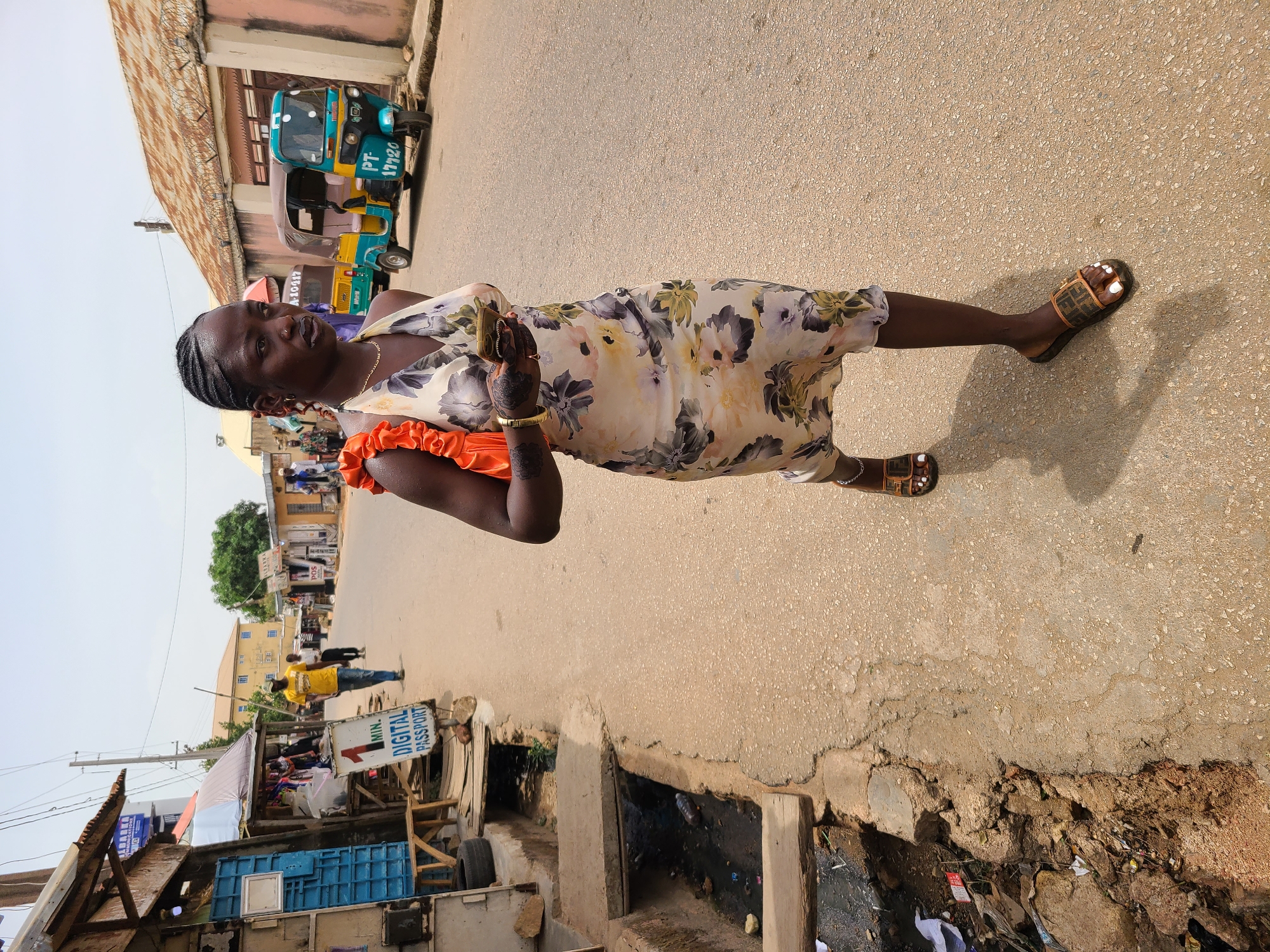 A medium shot captures a dark-skinned woman in Jos, Nigeria, standing on a paved road lined with buildings and drainage. The woman, positioned slightly off-center, wears a floral print dress with orange ruffled sleeves and flat sandals. Her hair is braided and pulled back. She holds a small object in her hands. A blue and yellow motorized tricycle is parked in the background. A sewage drain is in the foreground with visible litter. Several figures are seen in the background, including a man in a yellow shirt.