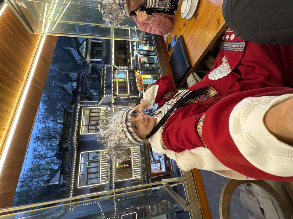 An individual is seated indoors at a wooden table, taking a selfie, and smiling at the camera. This person, of apparent European descent, is adorned in a red traditional Chinese-style robe with white trim, a fur-lined collar, and elaborate silver ornamentation, including a large, ornate silver headdress characteristic of Miao ethnic attire. Several dark braids with silver accents are visible over their left shoulder. A second individual, partially visible to the right, also wears a similar silver headdress and a pink puffy jacket, looking down. On the table between them rests a black folder, a smartphone, and a white dish with blue patterns.
The scene is set in Fenghuang Ancient Town, China, at dusk or night. Through a large window overlooking a river, traditional Chinese buildings with multi-tiered roofs and balconies are visible across the water. These buildings are extensively illuminated with white string lights and strip lighting along their edges and windows, creating a vibrant evening atmosphere. Distant hills with sparse trees are visible under a dark blue sky above the illuminated structures. The interior space features a warm, amber light strip running along the ceiling.