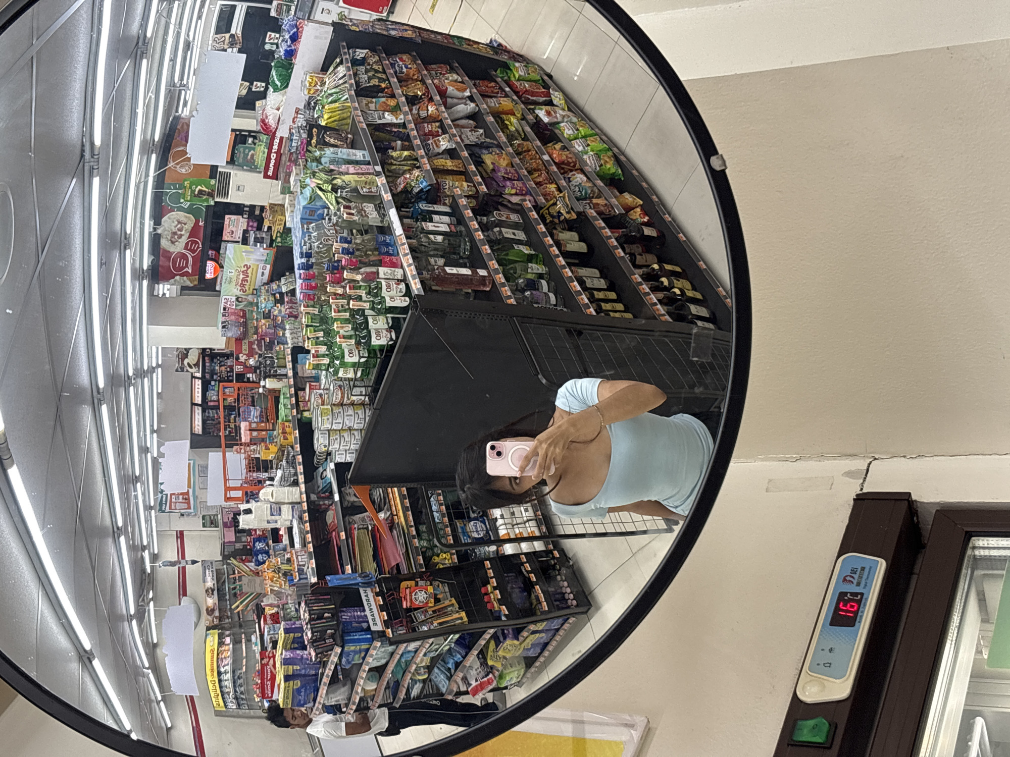 The image is a fisheye mirror reflection of the interior of a convenience store, likely a 7-Eleven, in Davao City, Philippines. In the foreground, a young woman is taking a selfie with a pink smartphone. She is wearing a light blue top. In the background, a man in a white t-shirt and dark pants stands near shelves stocked with various products. The store is well-lit with fluorescent lights and features aisles of merchandise, including beverages, snacks, and household items. A digital thermometer on the wall displays "16C." The store appears to be clean and organized.