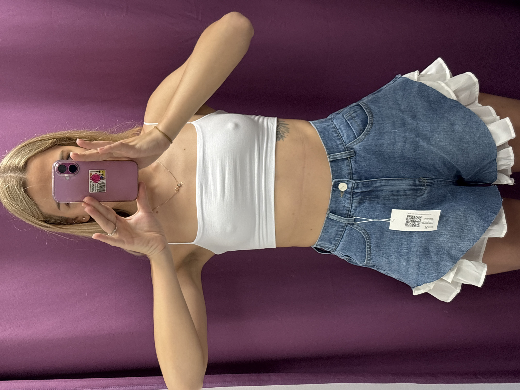 A woman is taking a selfie in a dressing room. She is wearing a white crop top and denim shorts with ruffled white trim. She is holding a pink smartphone in her hands, obscuring her face. The background is a purple curtain. The image does not provide any information about the location of the city of Krasnoyarsk, Russia.