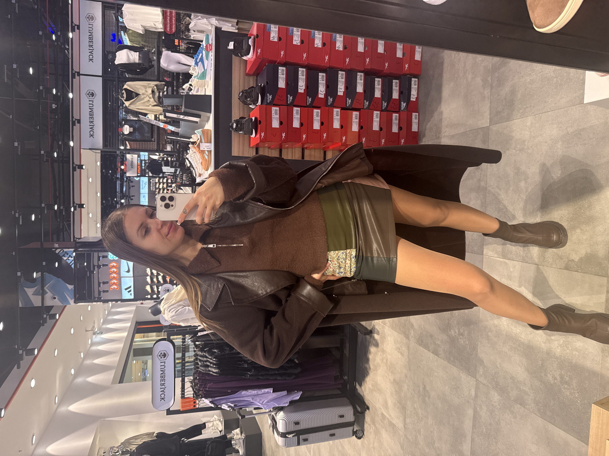A woman is taking a selfie in a shop, likely in Batumi, Georgia, based on the upside-down "Lumberjack" signs. She is wearing a brown jacket, a brown sweater, and a green skirt. She also has brown boots on. The shop appears to sell shoes, clothes, and luggage. Rows of shoe boxes are visible on shelves behind her. Clothes are hanging on racks nearby. The floor is gray tile.