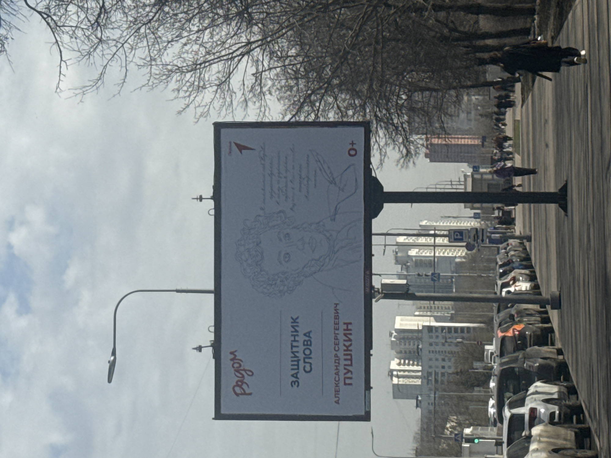 The image shows a street in Moscow, Russia, with a billboard promoting Alexander Pushkin, a famous Russian poet and writer. The billboard features a sketch of Pushkin's face and the words "Защитник слова" ("Defender of the word") and "Александр Сергеевич Пушкин" ("Alexander Sergeyevich Pushkin"). A street lamp and some tree branches are visible near the billboard. In the background, there are high-rise buildings and a line of parked cars along the street. People are walking along the sidewalk. The sky is overcast.