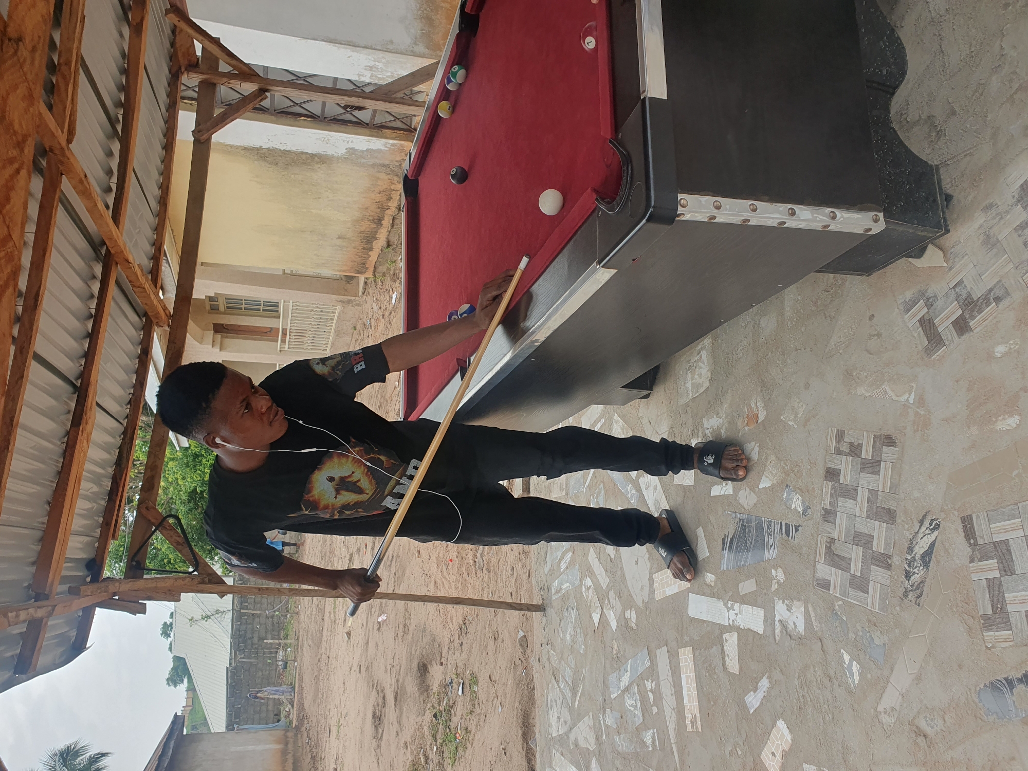 A young Black male stands to the left of a red billiards table, holding a cue stick in his right hand and aiming at a white ball. He is wearing a black t-shirt with a graphic print, black pants, and black slide sandals. White earbuds are in his ears. The billiards table is outdoors under a slatted wooden roof structure. The ground around the table is a mixture of sand and irregular mosaic tiles. In the background, there are buildings with light-colored walls and a partially visible fence. The sky is overcast. The overall scene appears to be in an outdoor recreational area, potentially a local gathering spot, in Lokoja, Nigeria.