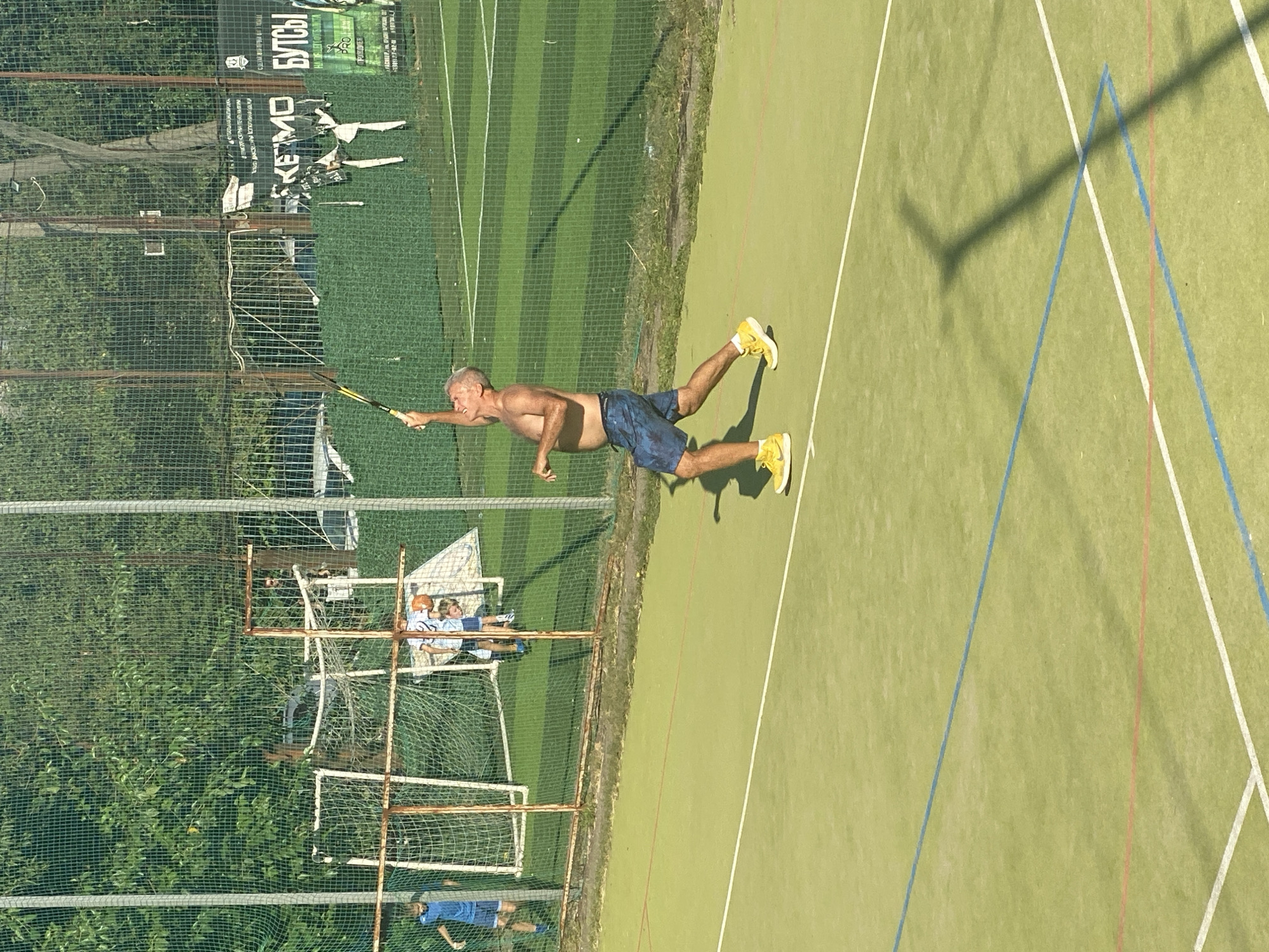 An older Caucasian man, with graying hair and a shirtless torso, is actively engaged in playing tennis on an outdoor court. He is positioned mid-swing, with his right arm extended upwards, holding a tennis racket as if to serve or hit a ball. He wears blue and black patterned shorts and bright yellow tennis shoes. The court surface is a vibrant green with distinct white and blue lines demarcating boundaries. In the background, several younger individuals in athletic wear are visible near a net and goalpost, suggesting a sports complex or park setting. The environment is outdoors with a fence and trees in the distance, indicating a sunny day. The image appears to be taken in the city of Krasnodar, Russia, as suggested by a visible sign in the background with Cyrillic text that reads "КЕЙМО БУТСЫ".