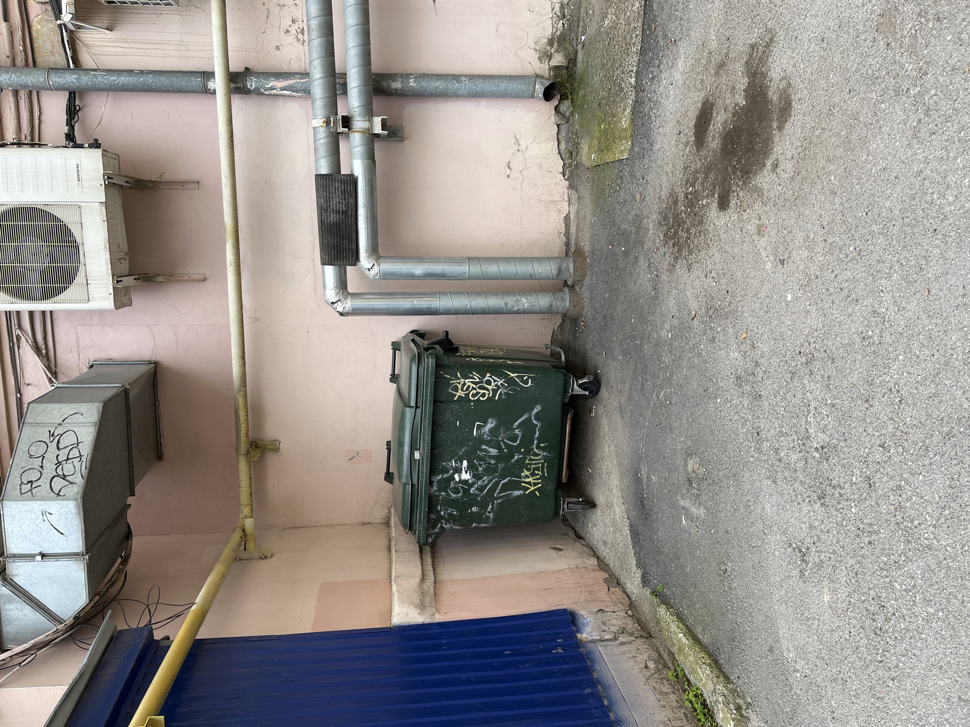 A green industrial dumpster with wheels is situated in an alleyway between two buildings. The dumpster is covered in white and yellow graffiti. To the left of the dumpster is a blue corrugated metal wall. To the right of the dumpster are several large grey pipes running horizontally and vertically along a light pink building. An air conditioning unit is mounted on the building above the pipes. The ground is paved with asphalt and shows signs of wear, including stains and small cracks. A metal ventilation system is visible above the pipes, also on the pink building.