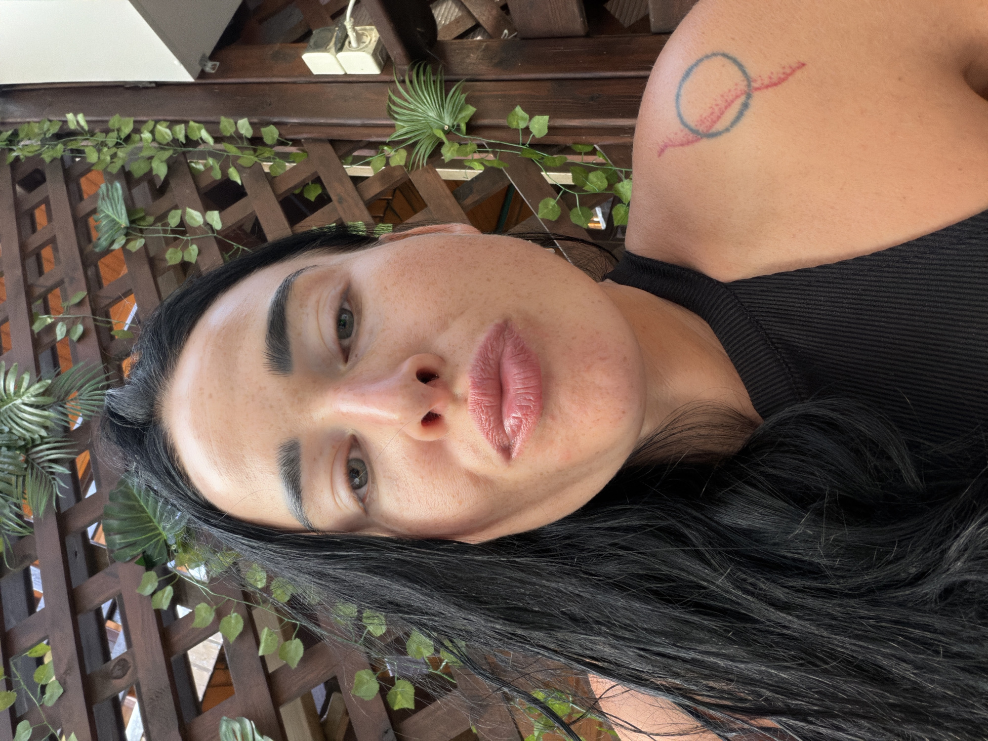 The image is a selfie taken by a woman with long, dark hair, freckles, and light-colored eyes. She is wearing a black tank top and has a tattoo on her right shoulder. The background appears to be an outdoor setting with a wooden lattice structure and hanging plants. The lighting suggests it is daytime. The image does not provide any specific information about the location being Sochi, Russia.