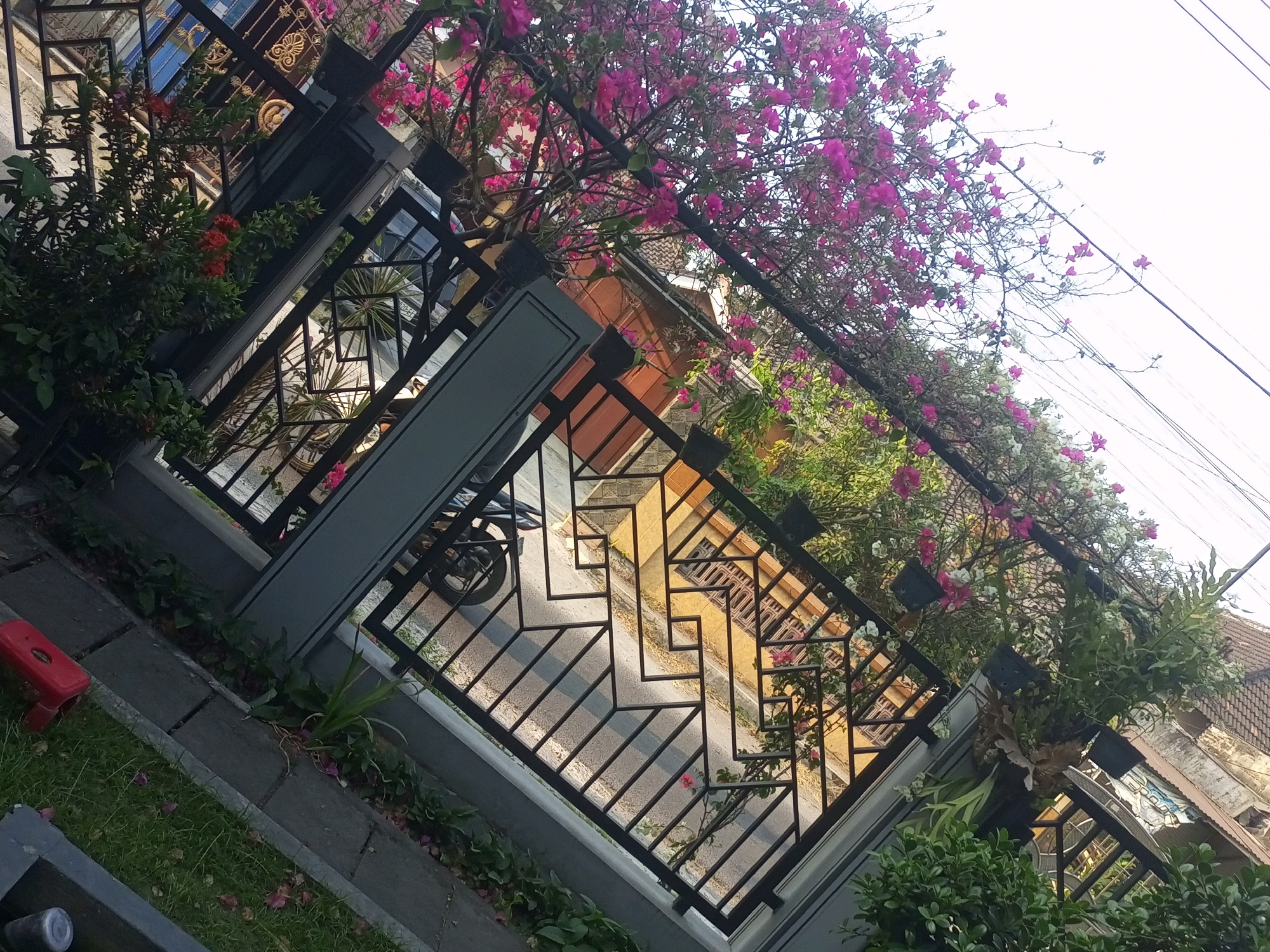 This image depicts a street-level view looking upwards towards a building with an ornate metal fence and flowering plants in Ngasem, Indonesia. The fence, made of black metal, features geometric patterns and is adorned with vibrant pink and white bougainvillea flowers that cascade over it. Behind the fence, a building with a tiled roof and windows is visible. The street below shows a paved surface and a motorcycle parked. The surrounding environment includes lush greenery and a cloudy sky.