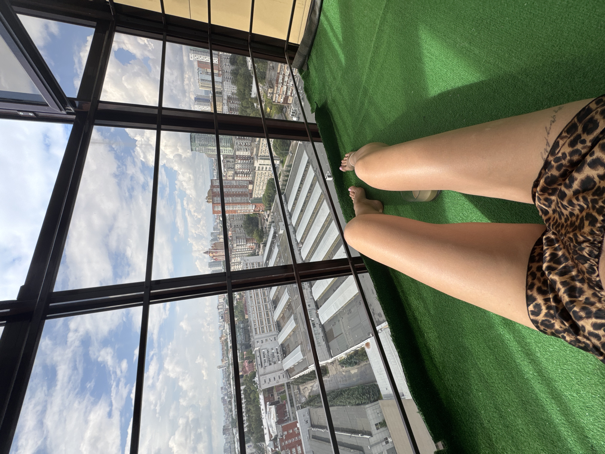 This image shows a first-person perspective of a person lying on a green, artificial turf surface on a balcony. The balcony has large, dark-framed windows offering a view of a city with many buildings, including some tall, modern structures and some older apartment buildings. The sky is cloudy with patches of blue. The person is wearing leopard print shorts and has bare legs. Their feet are visible, and one has a delicate anklet. A glass of what appears to be a light-colored beverage is placed on the turf near the person's legs. The urban landscape visible through the windows includes factories or industrial buildings with long, flat roofs in the foreground, transitioning to a denser residential area with more diverse architecture in the background. The overall impression is one of relaxation and enjoying the city view from a private space.
