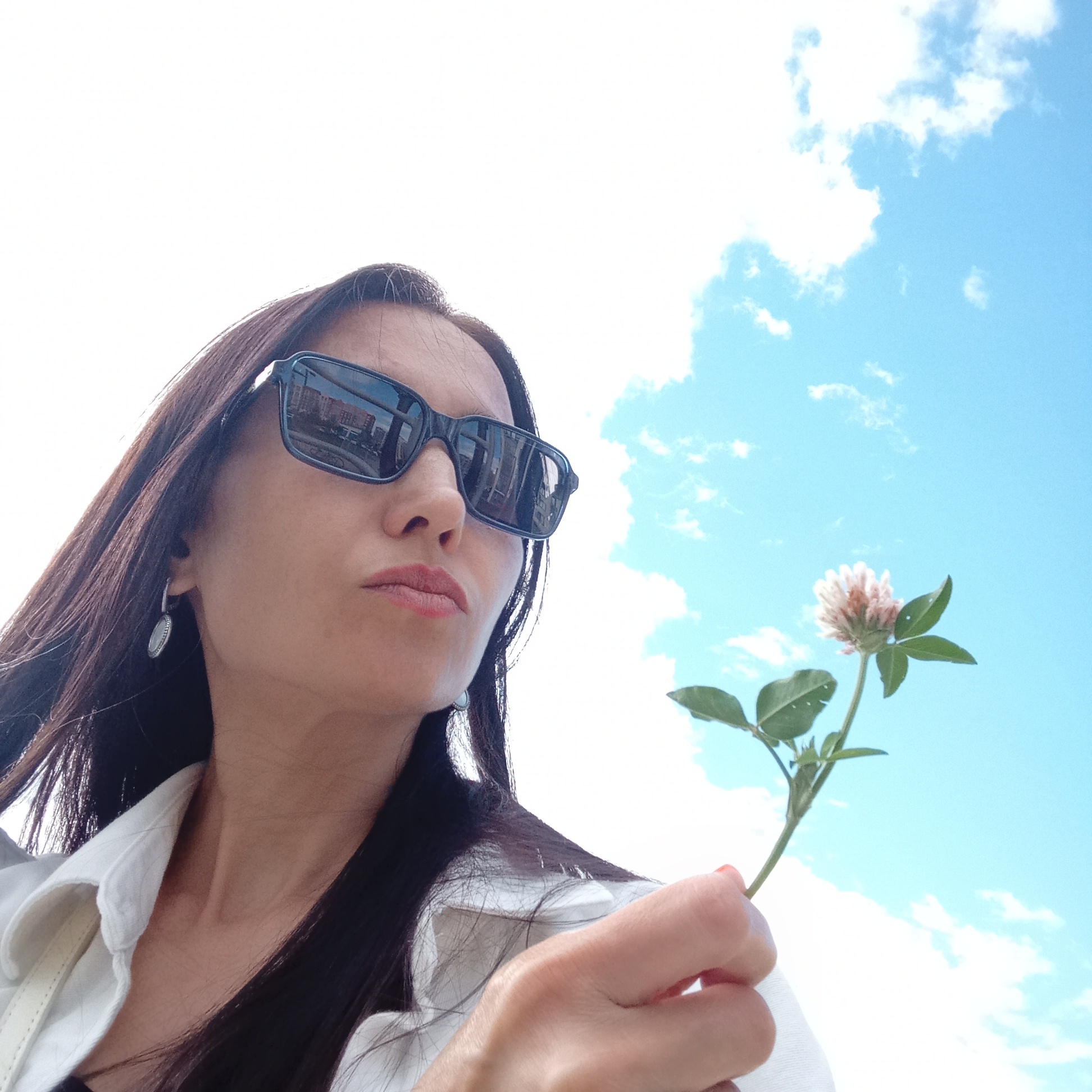 A woman wearing sunglasses holds a small clover flower against a bright blue sky with some white clouds. The reflection in her sunglasses shows buildings. The image appears to be a selfie taken outdoors in the city of Izhevsk, Russia.