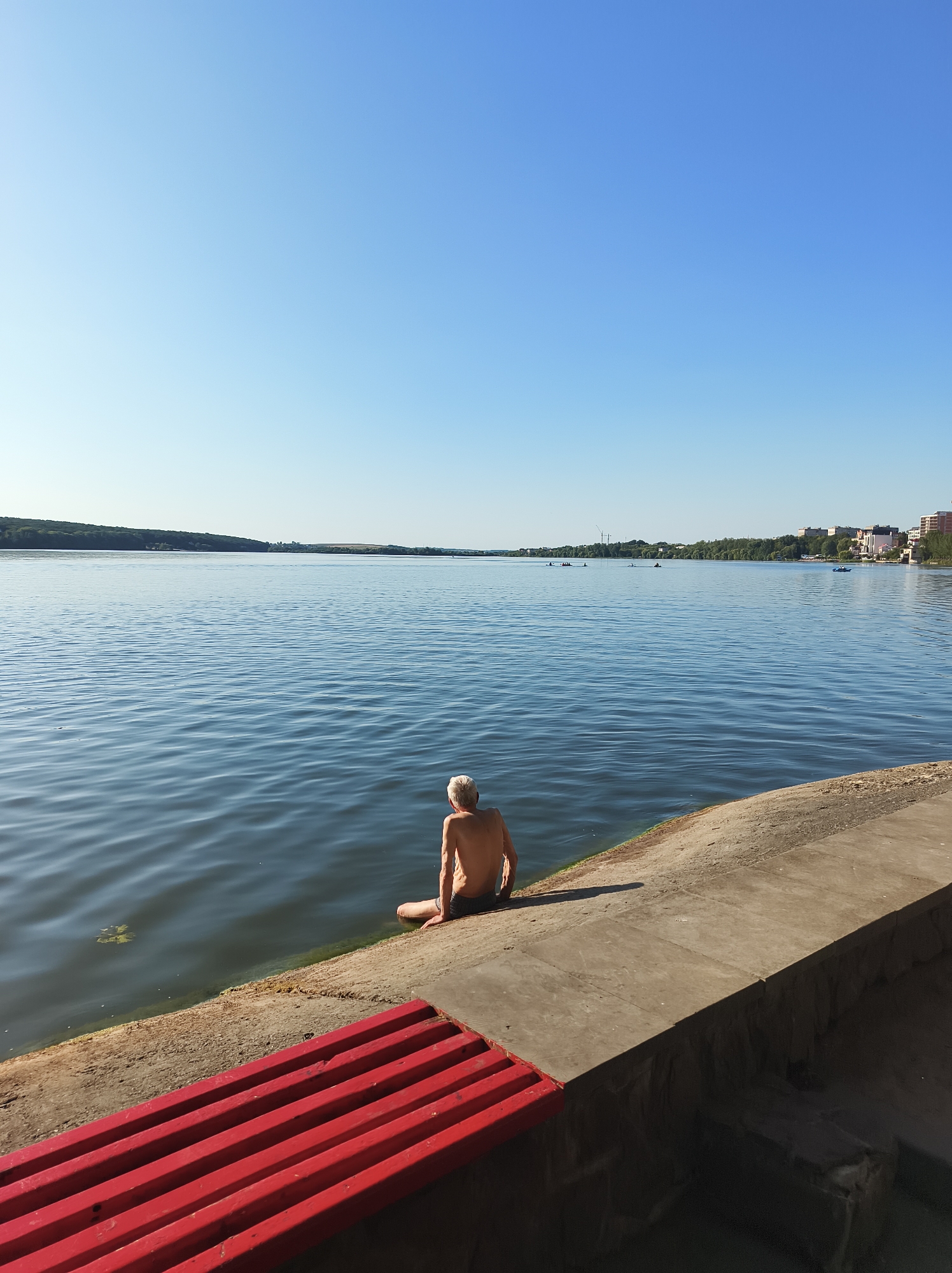 The image depicts a wide, calm lake under a clear blue sky. A bare-chested individual with white hair is seated on the concrete embankment, facing the water. In the foreground, a bright red wooden bench is visible. The background shows a distant tree-lined shore with some buildings and wind turbines. The scene appears to be a public waterfront area, possibly in the city of Ternopil, Ukraine, known for its prominent lake.