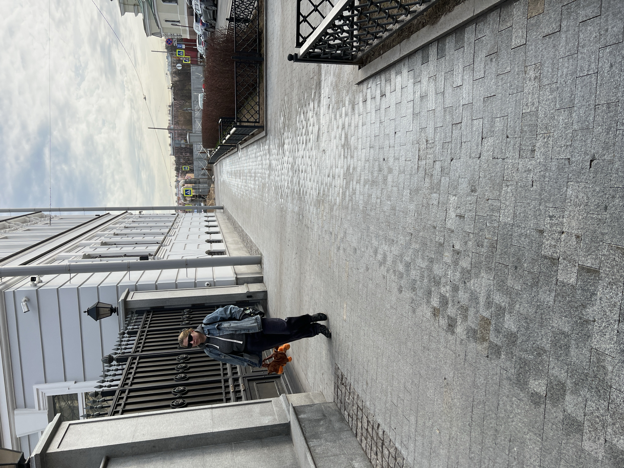 The image shows a woman standing on a gray, cobblestone sidewalk in Saint Petersburg, Russia. She is wearing a denim jacket, dark pants, and sunglasses, and she is holding a brown object. Behind her is a white building with a black wrought-iron gate. The sidewalk extends into the distance, bordered by a low black fence on one side and the building on the other. In the background, there are other buildings and a street with traffic signs. The sky is overcast.
