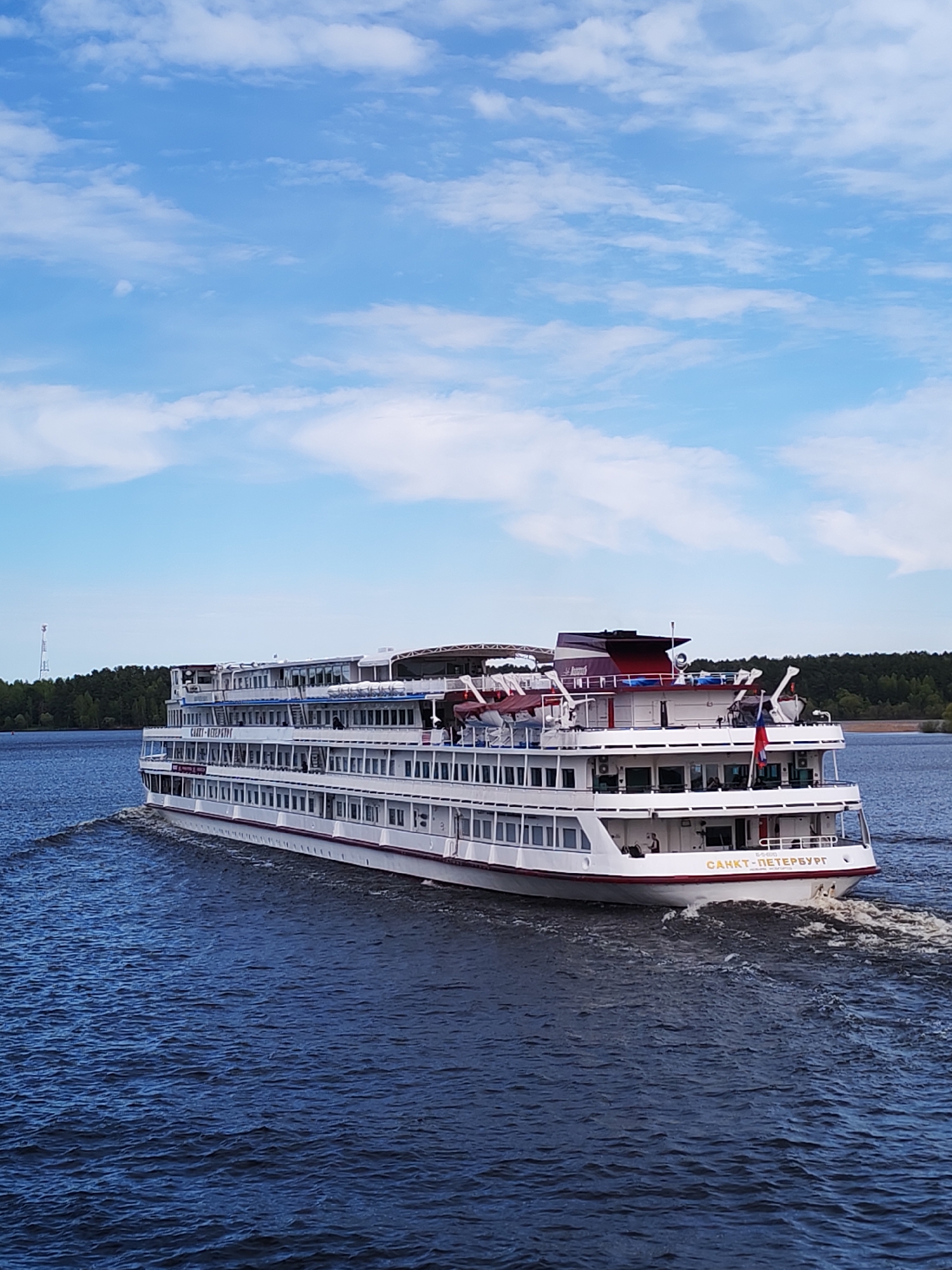 A large, white passenger cruise ship labeled "CAHKT-ПЕТЕРБУРГ" (St. Petersburg in Russian) on its stern is sailing on a body of water. The ship has multiple decks with numerous windows, a dark red and white structure on its top deck, and a small Russian flag is visible. The water is a deep blue and the ship is creating a noticeable wake. In the background, there is a strip of green trees along the shoreline and a tall, narrow antenna tower. The sky is blue with scattered white clouds, suggesting a clear day. The location is most likely the city of Myshkin, Russia.