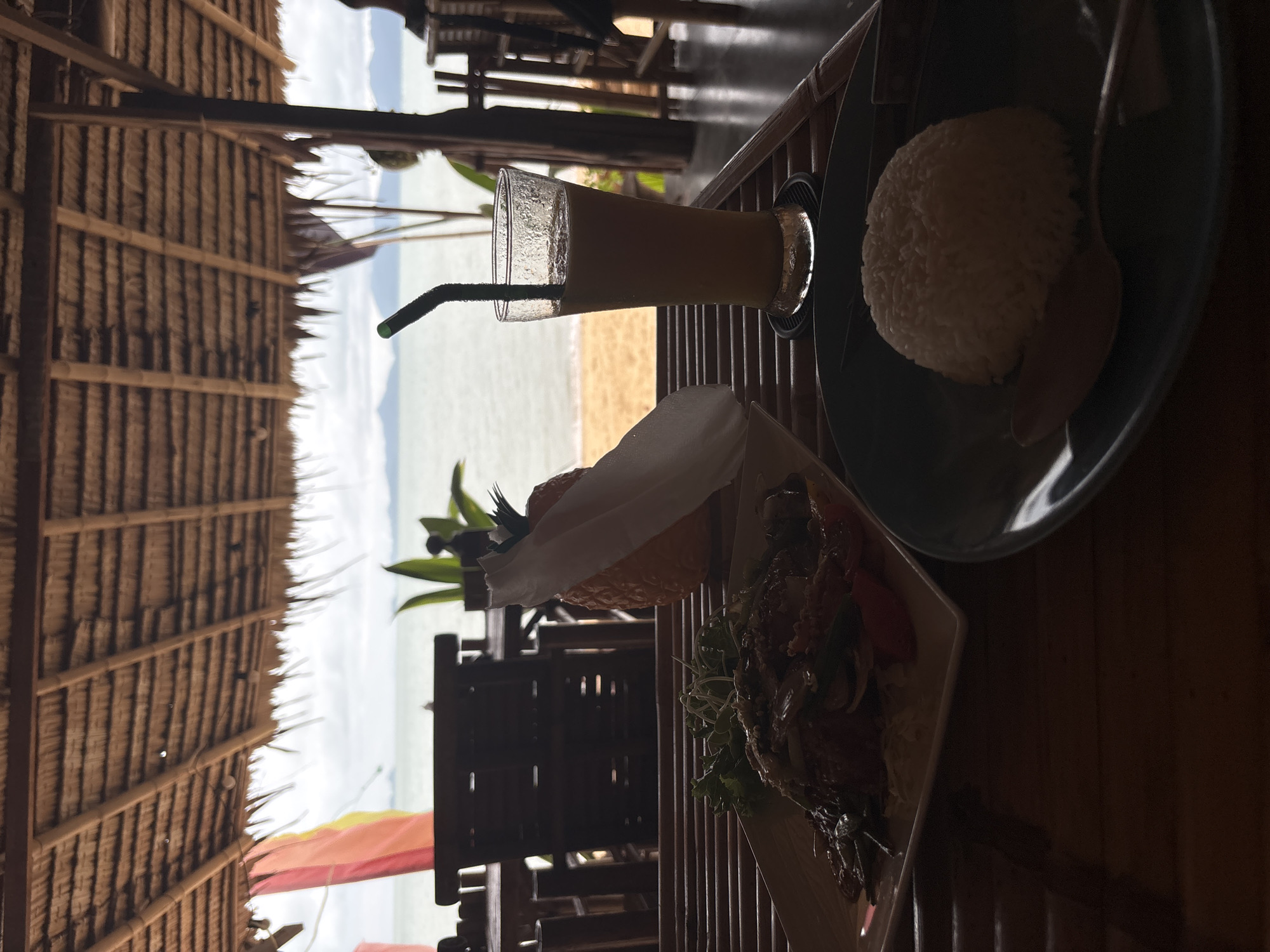 From an elevated perspective within a structure with a thatched roof, a table is laid with food and a beverage in Baan Bo Phut, Thailand. The scene includes a glass of orange-colored juice with a green straw, a plate of rice, and another dish that appears to contain salad or vegetables with a wrapped piece of fruit, possibly an orange. Outside, the beach and sea are visible in the background.