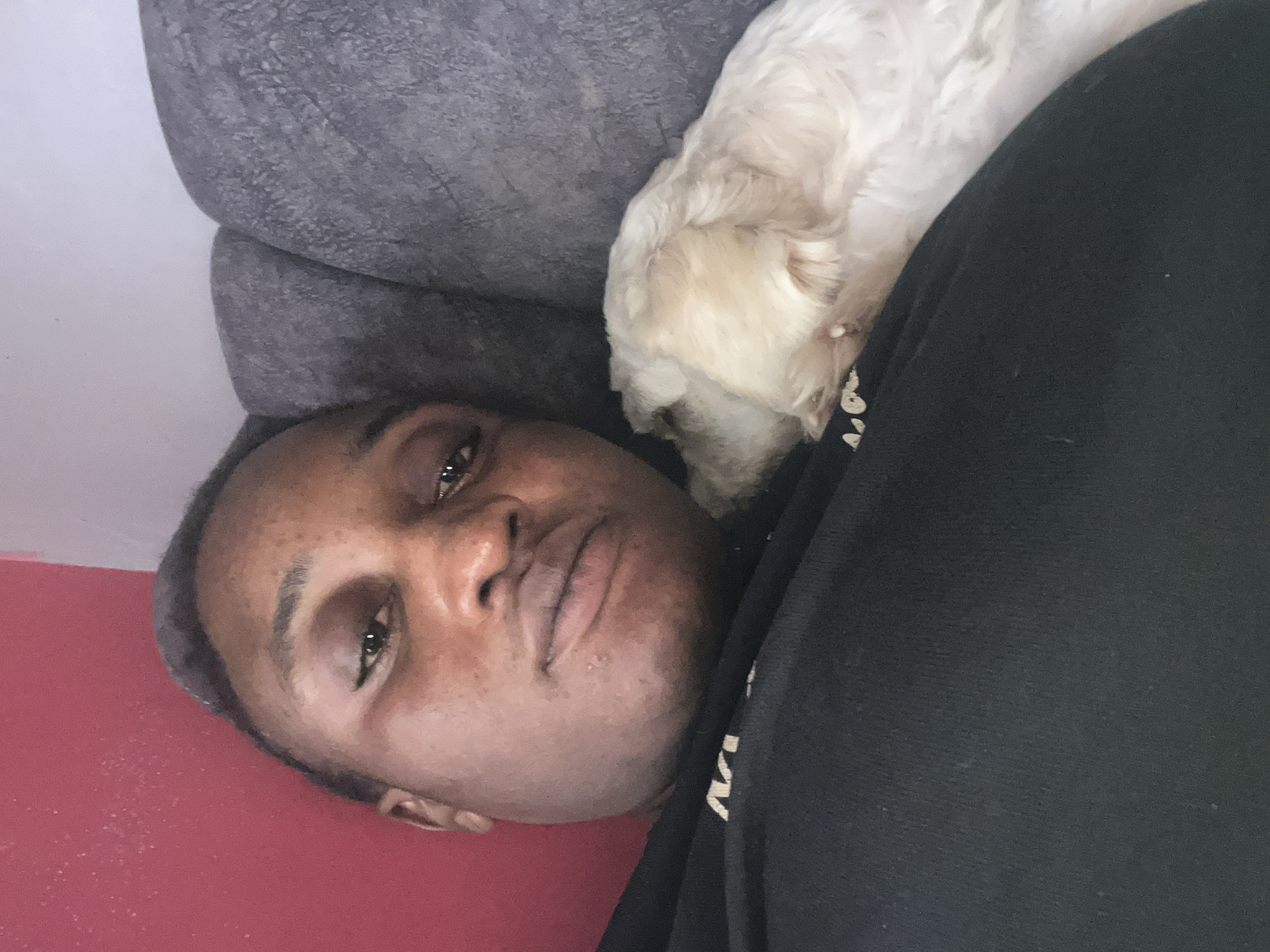 In Barakin Lerawi, Nigeria, a close-up selfie captures a person lying down with a small, light-colored dog resting on their chest. The person's face, partially obscured, is looking towards the camera, while the dog is facing away. The background features a textured grey sofa and a segment of a red wall. The person is wearing a black top, and the overall lighting suggests an indoor setting.