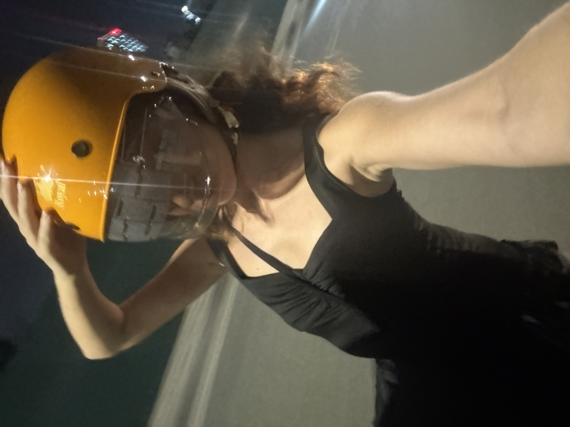 This is a first-person perspective photo of a person wearing a bright yellow full-face helmet with a clear visor. The person is wearing a black tank top. The helmet is held up to their head with their left hand. The background is blurry and appears to be an outdoor setting at night. There are some lights visible in the upper background, suggesting artificial lighting from buildings or vehicles. The overall impression is that the person is either about to ride or has just finished riding a motorcycle or similar vehicle in an urban environment, likely Hà Nội, Vietnam, given the location context. The lighting suggests it's evening or night.