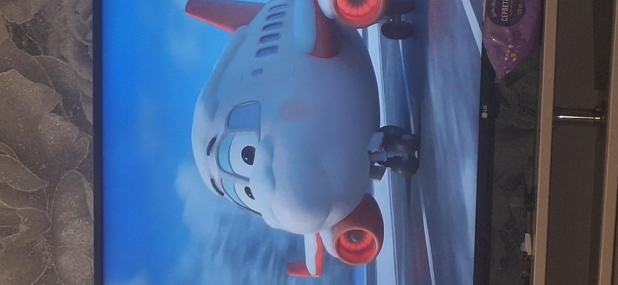 This is a close-up, medium shot of a cartoon airplane with a smiling face, positioned on a tarmac. The airplane is white with red accents on its wings and tail. Its large, round engines are visible, as are its landing gear. The background is a blurred blue sky with some clouds. Below the screen, a shelf is visible with various objects, including a purple package with Cyrillic writing and an LG logo on the television. The overall scene suggests a children's animation or game. There is no indication of the location being Bila Tserkva, Ukraine.