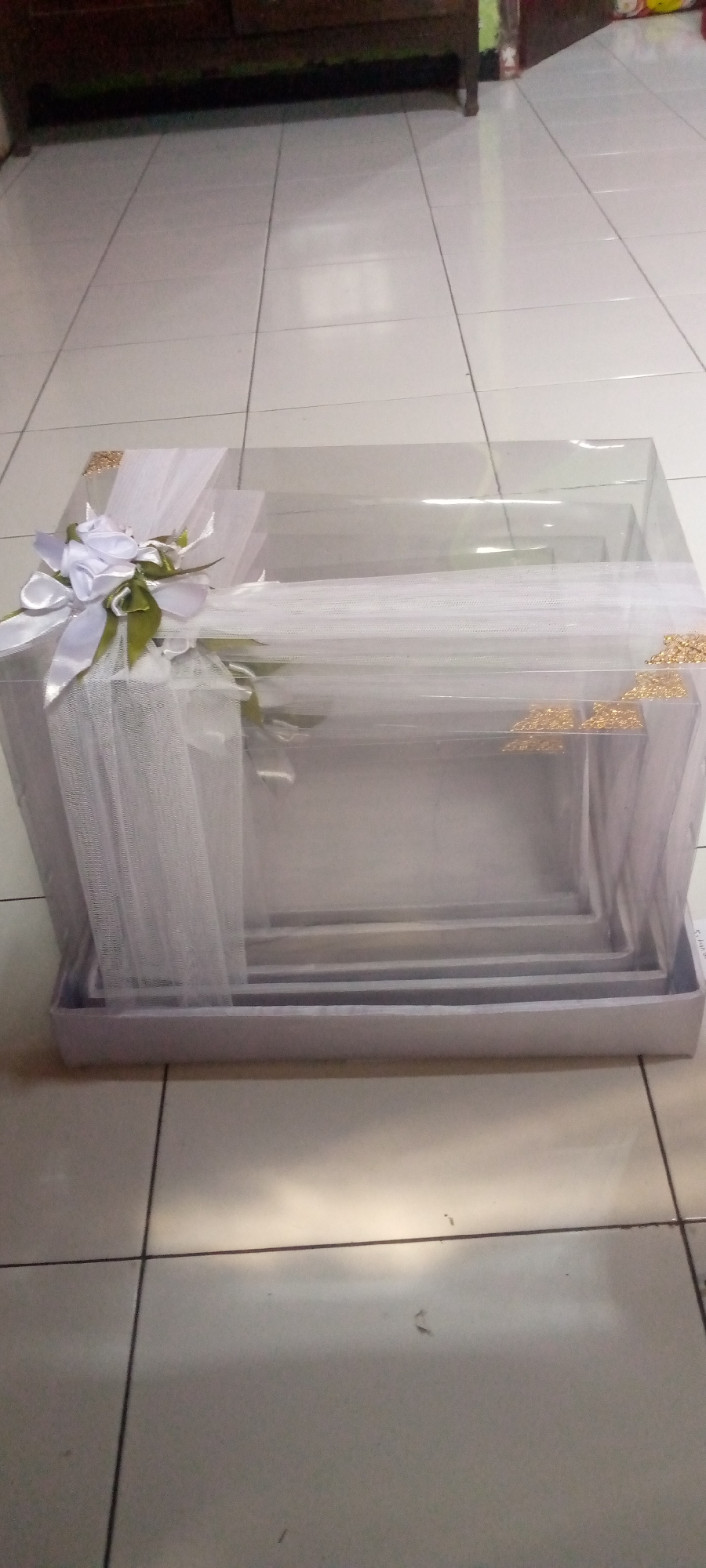 An overhead view of a clear plastic gift box is presented. The box is layered with multiple, smaller, clear plastic trays nested inside one another. White tulle fabric is draped over the top of the box, secured with a white ribbon bow and artificial green leaves on the left side. Gold decorative corner pieces adorn the edges of the upper box. The gift box rests on a light purple base. The background shows a tiled floor with dark grout lines. A dark wooden piece of furniture is visible in the upper portion of the image. The scene is indoors.