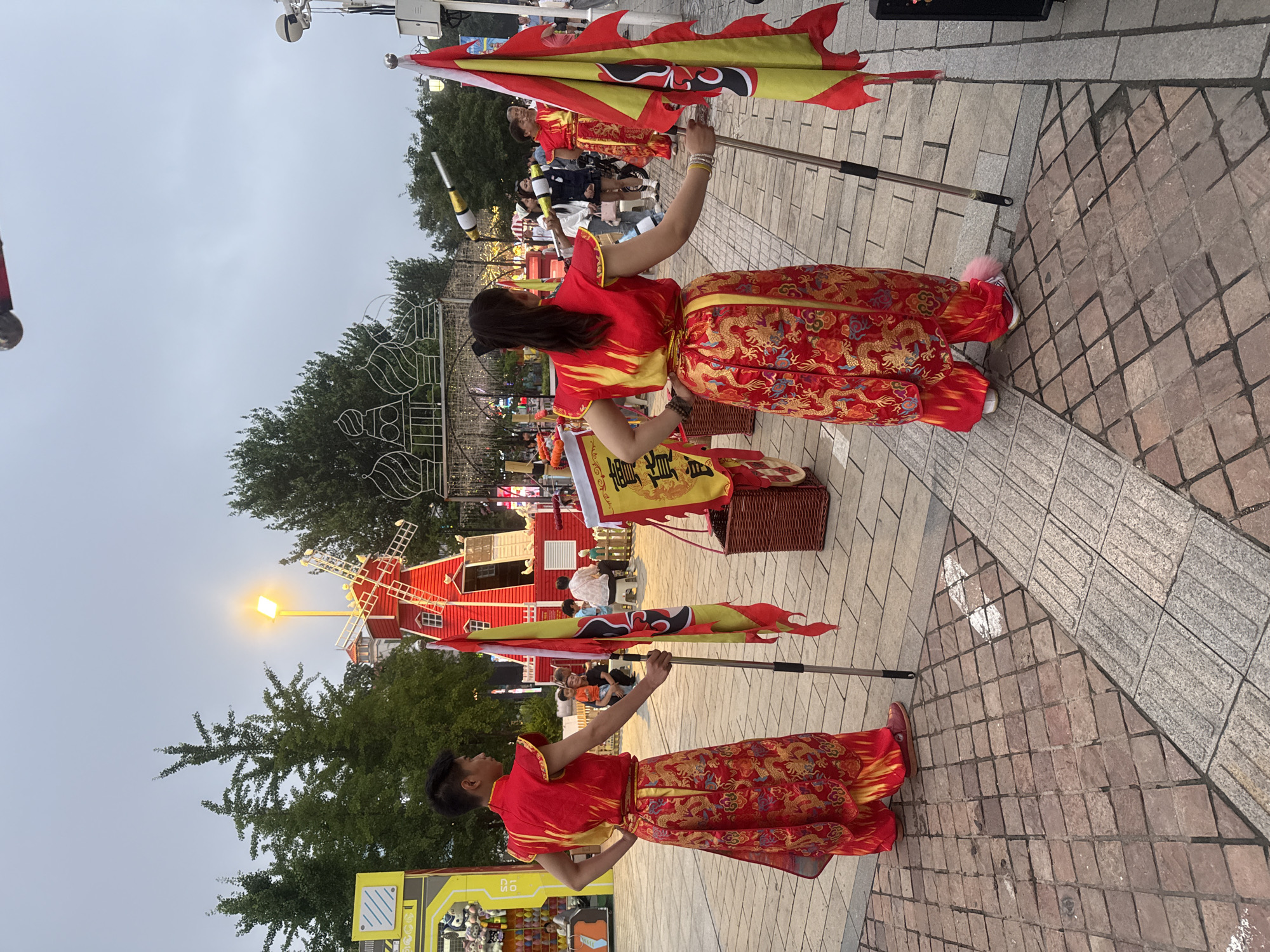 The image shows two individuals in traditional Chinese attire, performing in an outdoor setting. They are both wearing red and gold robes with intricate dragon patterns, and holding large, colorful flags on poles. The flags are red and yellow with black and gold accents. The ground is paved with cobblestones. In the background, there's a red miniature windmill, trees, and some people watching. The sky is overcast, suggesting it might be late afternoon or early evening. The overall atmosphere seems festive, possibly a cultural performance or parade. There are no clearly legible signs or text visible in the image. The location is identified as Qinhuangdao, China, which provides a cultural context for the attire and performance style.