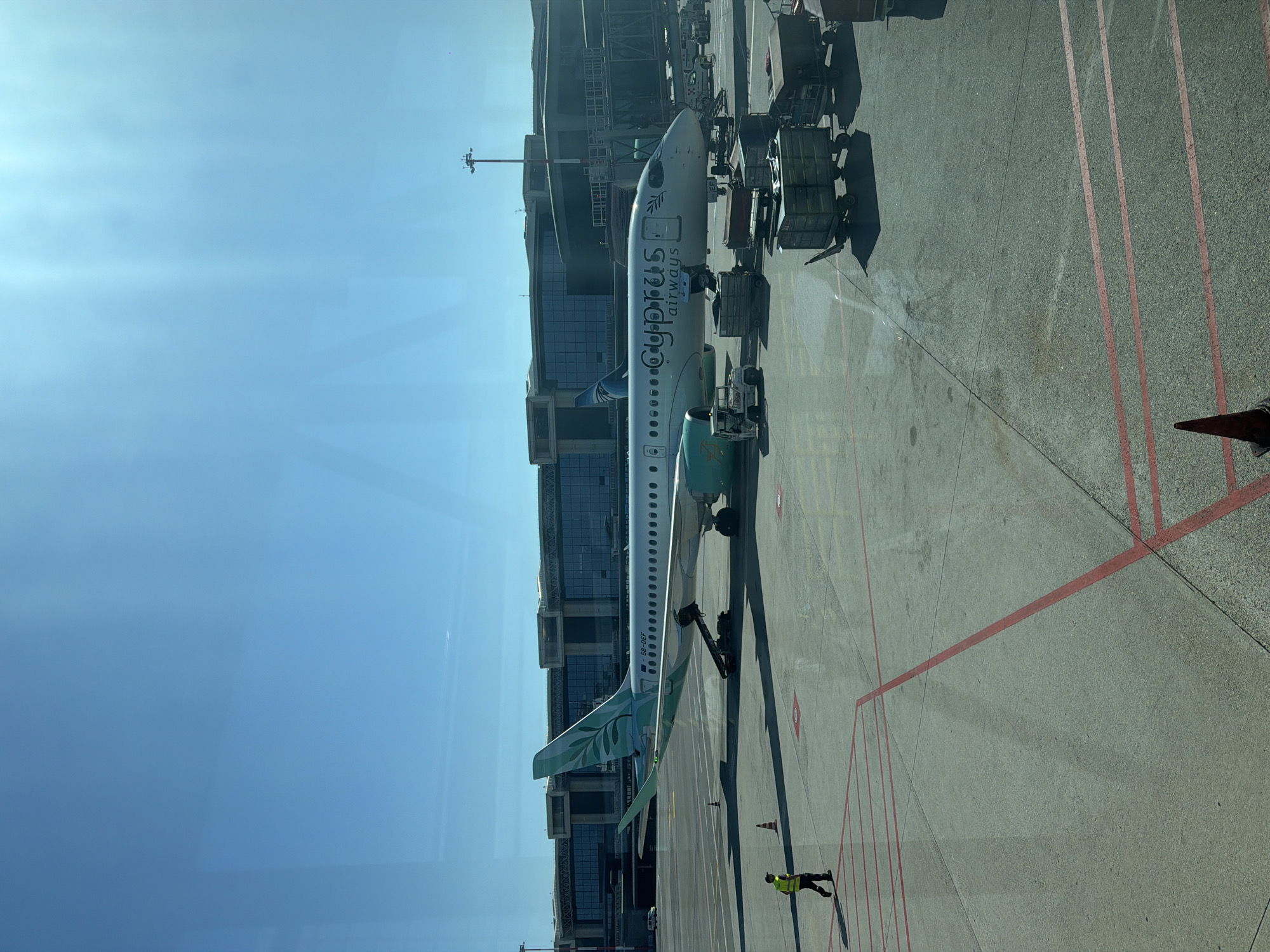 The image shows an airplane parked at an airport terminal. The aircraft, operated by Cyprus Airways, is predominantly white with turquoise accents on its tail and fuselage. The Cyprus Airways logo is visible on the side of the plane. Several service vehicles and luggage carts are positioned around the aircraft, suggesting ongoing ground operations. A person wearing a high-visibility vest is walking on the tarmac in the foreground. In the background, a modern airport terminal building with large glass windows and elevated walkways is visible, along with lighting poles. The scene is set on a sunny day, with a clear blue sky dominating the upper portion of the image. This scene is located at Milan Malpensa Airport (MXP) in Ferno, Italy.